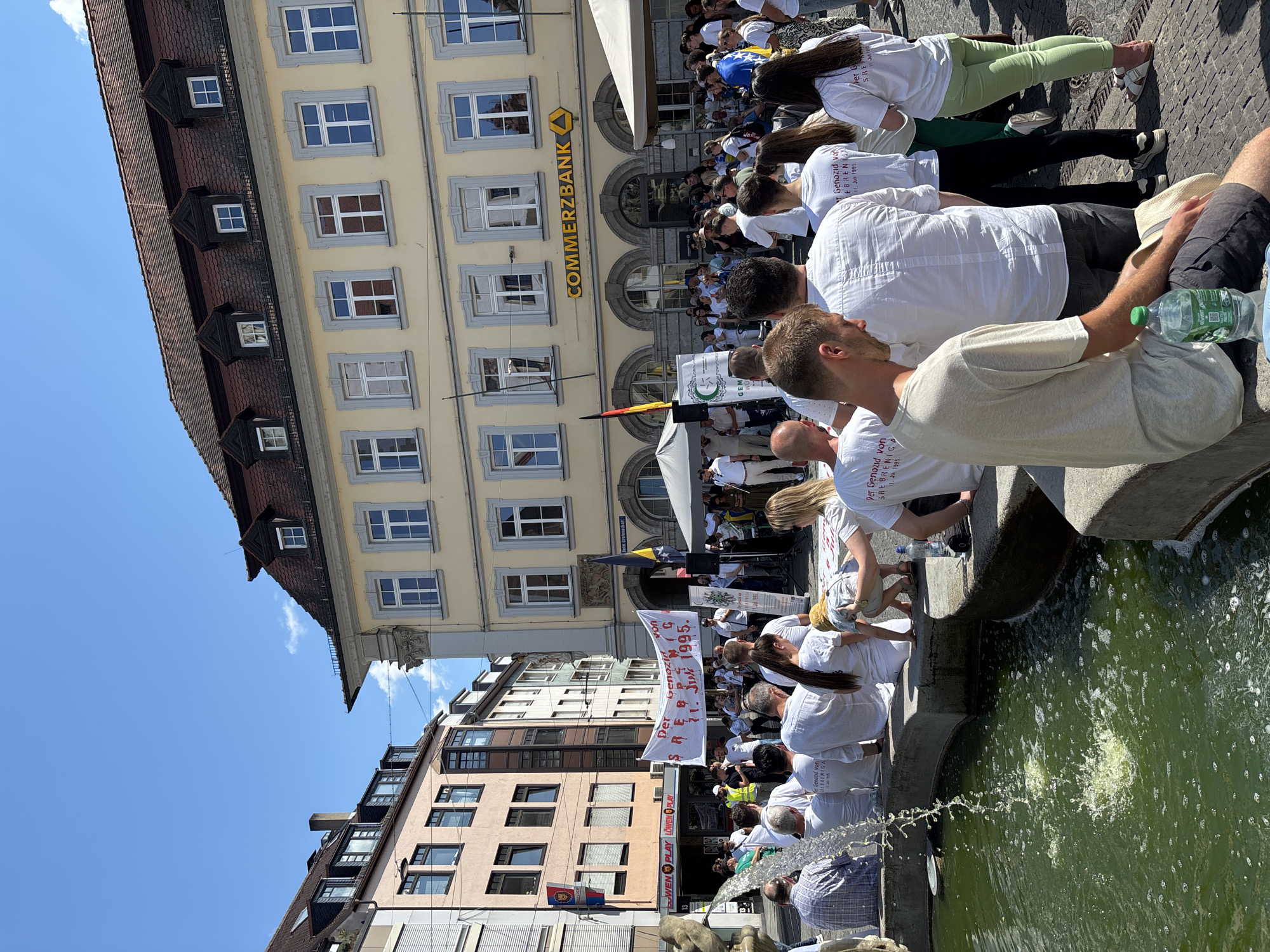 This is a medium-shot, eye-level photograph taken on a sunny day, capturing an outdoor gathering in Würzburg, Germany. The focal point of the image is a group of people, many wearing white t-shirts, clustered around a fountain. The t-shirts bear text in a foreign language, indicating a commemoration or protest. A large banner with similar text is held aloft by some individuals in the crowd.
In the background, a light yellow, multi-story building with classical architectural elements and numerous windows dominates the scene. A prominent sign on this building reads "COMMERZBANK". Adjacent buildings, typical of a European city center, are also visible, with shops and storefronts at street level. The sky is clear blue with a few wispy clouds.
The fountain in the foreground is made of concrete, with water cascading into a green-tinted pool. Several individuals are seated on the edge of the fountain, facing the central gathering. The overall atmosphere appears to be one of solemn observance or remembrance. The presence of flags, a stage-like setup with microphones, and the concentrated grouping of people suggest an organized event. The specific location in Würzburg appears to be a public square.