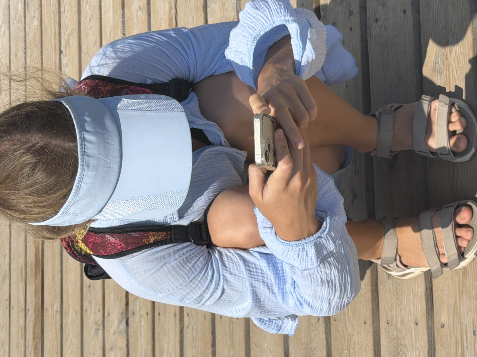 A person wearing a light blue long-sleeved shirt, a light blue visor, and beige sandals is crouching on a wooden boardwalk. The person is holding a silver smartphone in their hands. A backpack with a patterned strap is visible on their back. The scene appears to be outdoors in a sunny environment, likely a tourist area given the boardwalk and the person's attire. The lighting suggests it is daytime. The background consists of horizontally laid wooden planks.