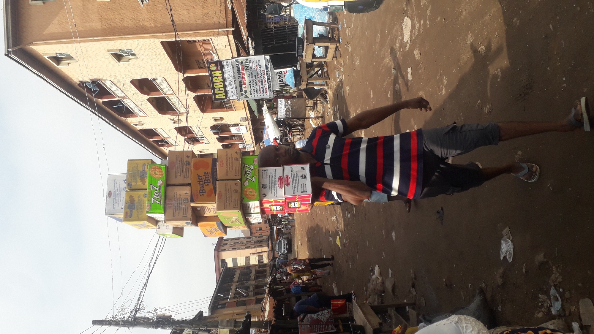 A man carries a tall stack of cardboard boxes on his head in a bustling outdoor market in Onitsha, Nigeria. The man is wearing a striped t-shirt and dark shorts. The boxes appear to be food products, with labels visible for "Butter Bite" and "7 to 7". In the background, there are market stalls, buildings, and other people navigating the crowded dirt pathway. The scene captures the vibrancy and commercial activity typical of Nigerian markets.
