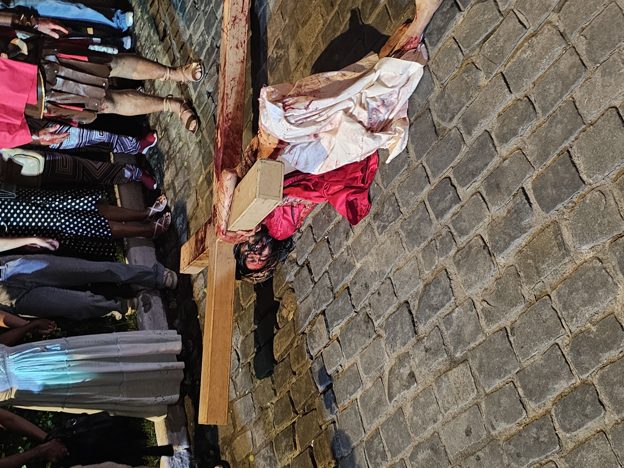 The image shows a re-enactment of the crucifixion of Jesus Christ on a cobblestone street in Armação dos Búzios, Brazil. A person portraying Jesus lies face down next to a wooden cross, wearing a crown of thorns and bloodied garments. Roman soldier impersonators are lying to the side, along with other people.