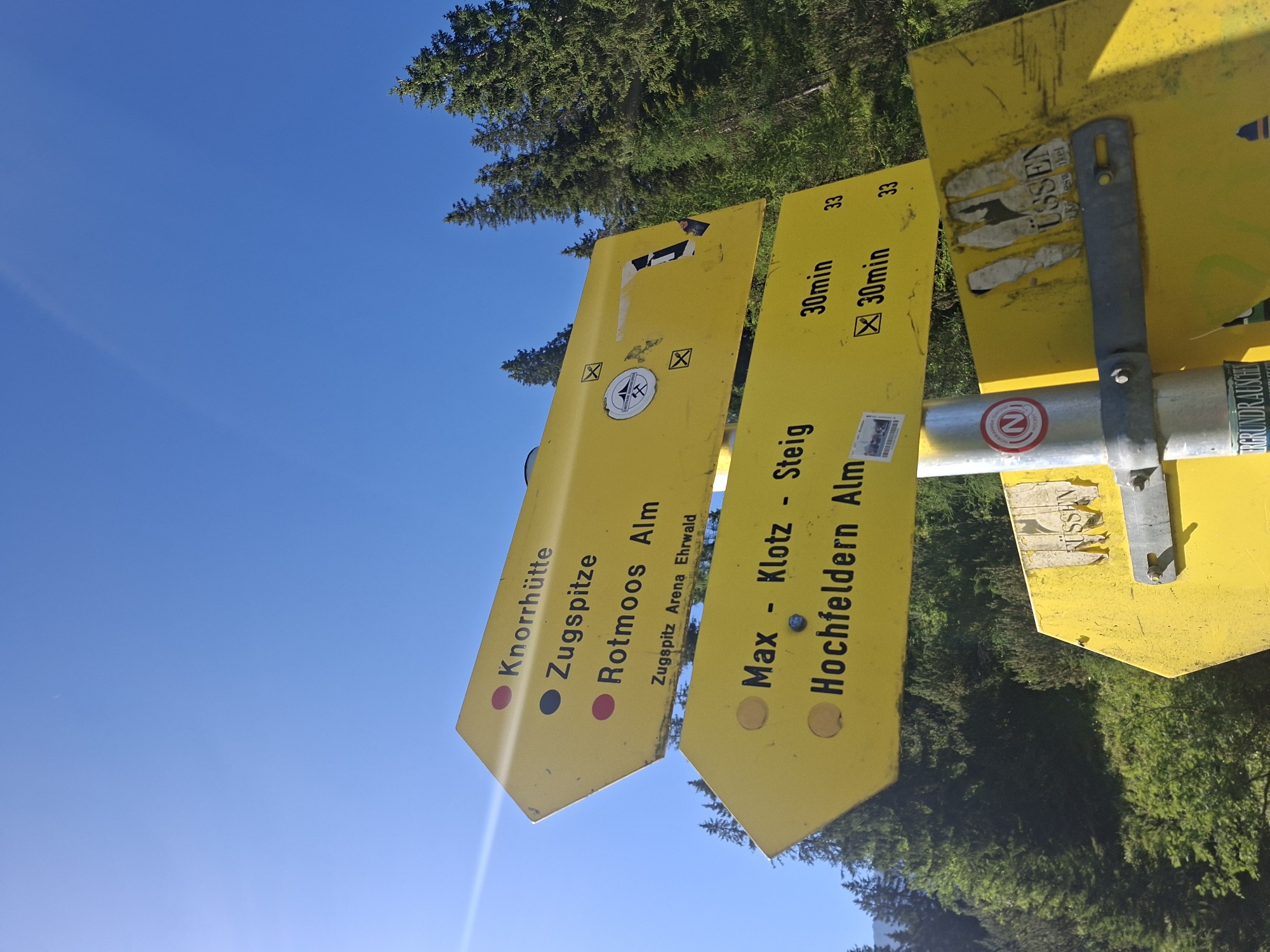 The image depicts a yellow hiking signpost with multiple directional arrows against a clear blue sky and a backdrop of a dense evergreen forest. The signpost, mounted on a metal pole, displays directions to various locations. The upper arrow points to "Knorrhütte," "Zugspitze," and "Rotmoos Alm," with "Zugspitz Arena Ehrwald" written below. The middle arrow indicates "Max - Klotz - Steig" and "Hochfeldern Alm." Time estimates for reaching these destinations are provided: "30min" for "Max - Klotz - Steig" and "30min" for "Hochfeldern Alm." The lower section of the signpost shows further directions, with one arrow pointing to "TÜSSEN" with some faded text beneath it. There are several stickers and markings on the signs, including a circular emblem with an "N" and some faded print. The environment is mountainous and forested, suggesting a trail or hiking area. The scene is captured in daylight, with sunlight glinting off the signpost.