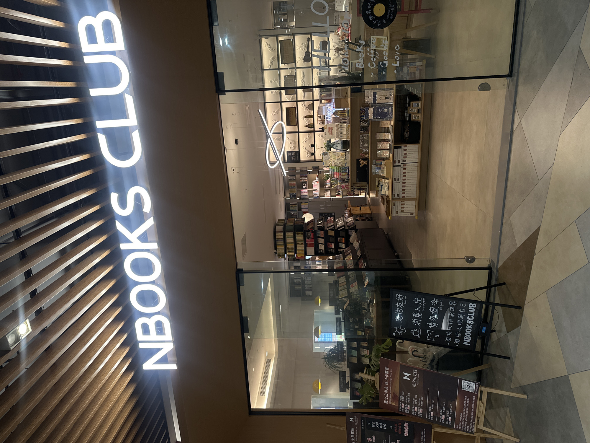 The image shows the exterior of a bookstore named "NBOOKS CLUB" located in the city of Chongqing High-tech Zone, China. The bookstore's name is displayed prominently in large white illuminated letters across the top of the entrance. The entrance features large glass doors and windows, offering a view into the interior.
Inside the bookstore, shelves are filled with books, and there are display cases with various items, including stationery, books, and decorative objects. The interior design appears modern, with wooden accents on the ceiling and sleek lighting fixtures. Several signs are visible, including a stand with information about membership recharge prices and a blackboard with handwritten text and drawings. One of the signs features the text "NBOOKS CLUB" and "南山书店" (Nanshan Bookstore). Another sign, a blackboard, has the text "宠物友好" (Pet Friendly), "请勿喧哗" (Please be quiet), "阅读以探索世界" (Read to explore the world), and "NBOOKS CLUB".
In the foreground, on the left side, is a wooden easel holding a menu-like board with Chinese text, including pricing information and operating hours. On the right side, a partial view of a red chair is visible through the glass. The flooring in front of the store is a light-colored tiled surface with a geometric pattern. The overall scene depicts a well-lit and organized bookstore environment.