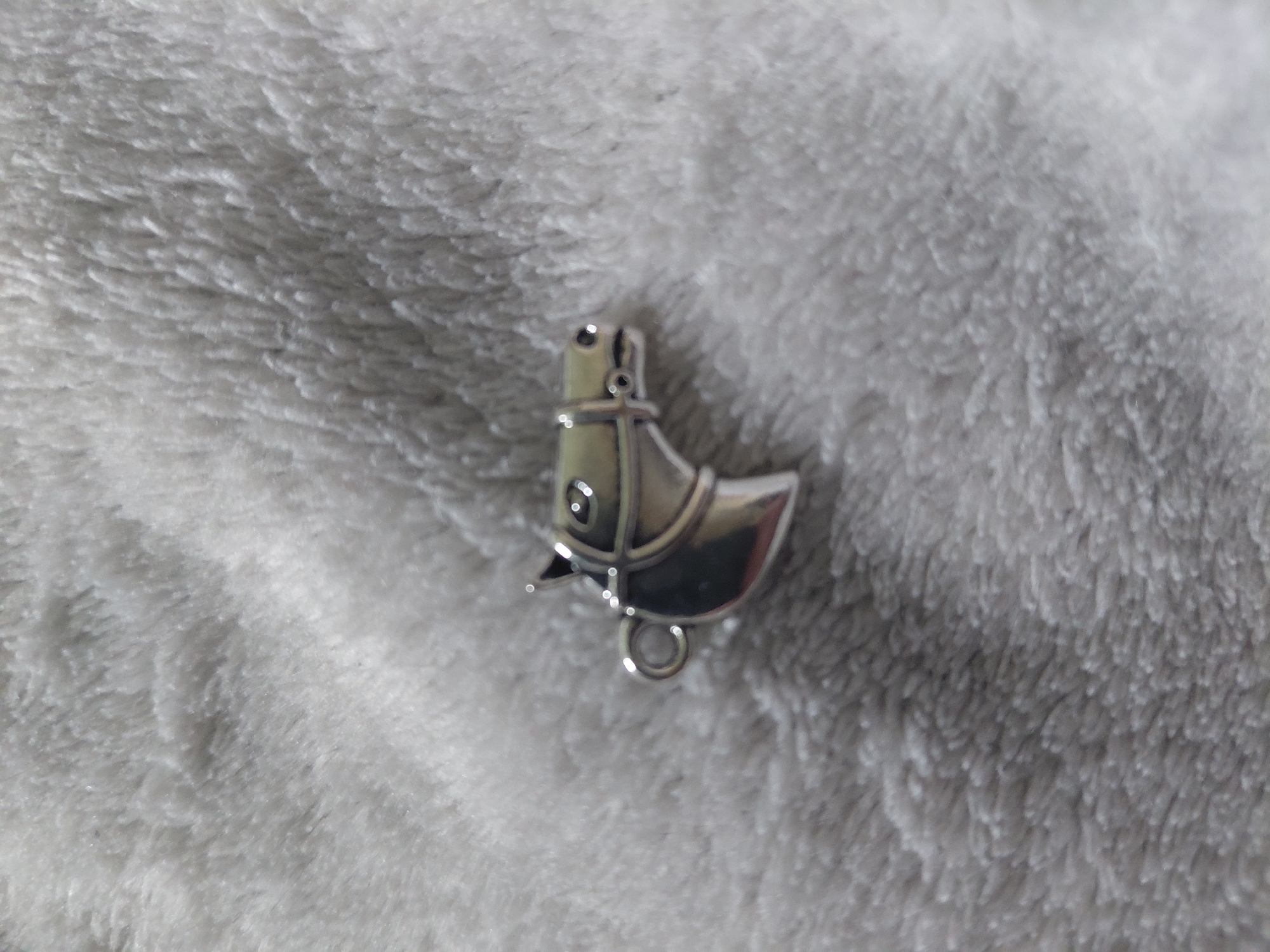 The image displays a small, silver-toned metallic charm shaped like a stylized horse's head, oriented to the right. The charm exhibits a polished, reflective surface with distinct highlights. Features include a prominent ear, an oval eye with a dark pupil, and a defined muzzle with subtle nostril depressions. A series of raised lines across the forehead and muzzle represent a bridle or headstall. A circular loop, or bail, is present at the rear base of the head, intended for attachment. The object rests centrally on a light grey, deeply textured fabric surface, likely faux fur or a plush textile. The fabric's fibers create a soft, uneven texture with varying shadows and light reflections. The background beyond the charm is softly blurred, indicating a shallow depth of field, with the object being the primary point of focus. No other subjects, objects, or actions are discernible in the scene.