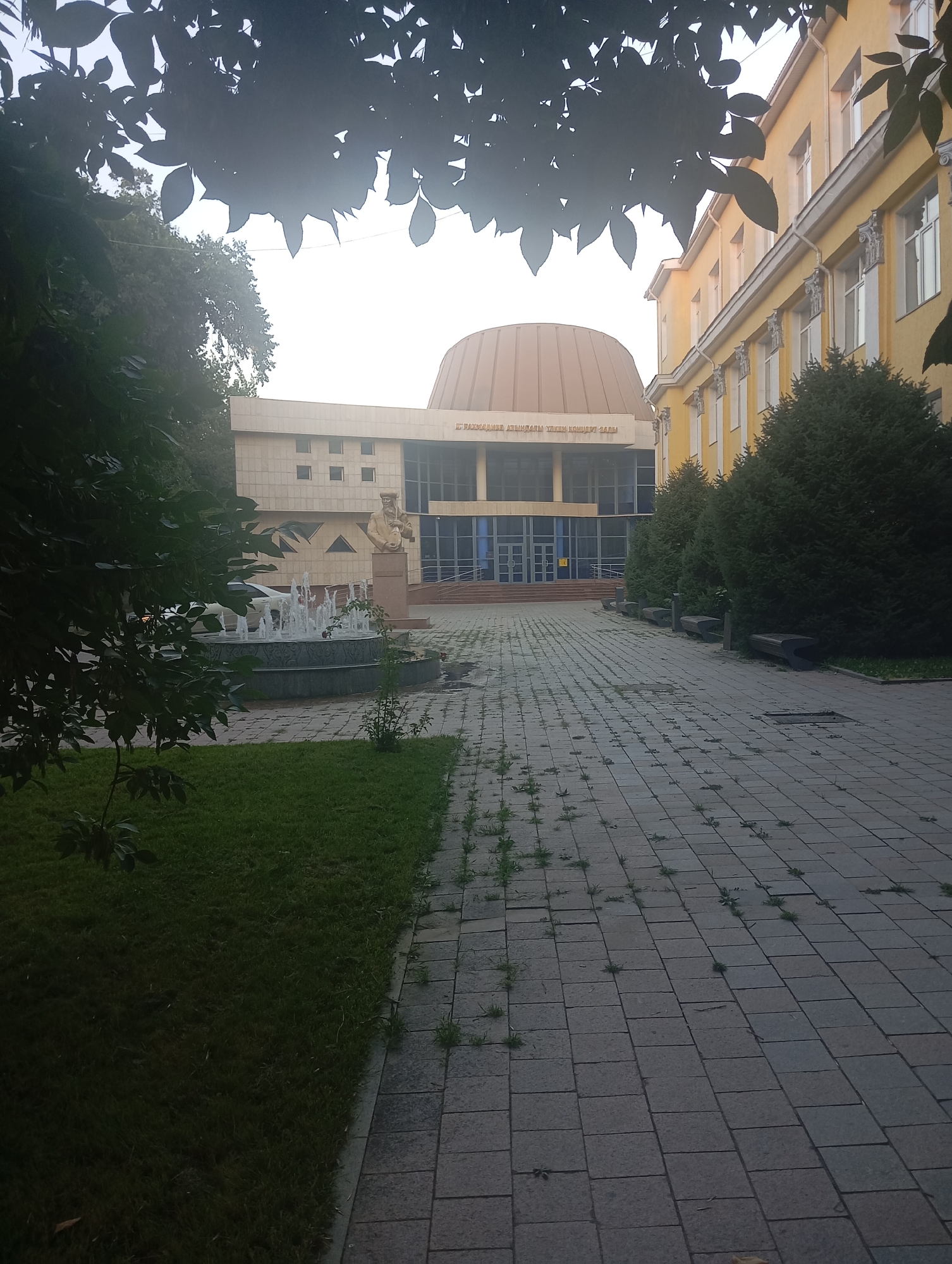 The image depicts an outdoor scene in front of a building with a distinctive domed roof, situated in Almaty, Kazakhstan. The building appears to be a cultural or educational institution, possibly a concert hall, as indicated by text visible on the facade. To the right of the domed structure, a multi-story yellow building with numerous windows is visible. In the foreground, a paved walkway made of square tiles leads towards the building. To the left of the walkway is a patch of grass and a circular fountain with water jets. A statue of a man in traditional attire stands near the fountain and the building entrance. Trees and foliage frame the scene, casting shadows and partially obscuring the view of the sky and the tops of the buildings. The overall impression is one of an institutional or public space, likely in an urban setting.