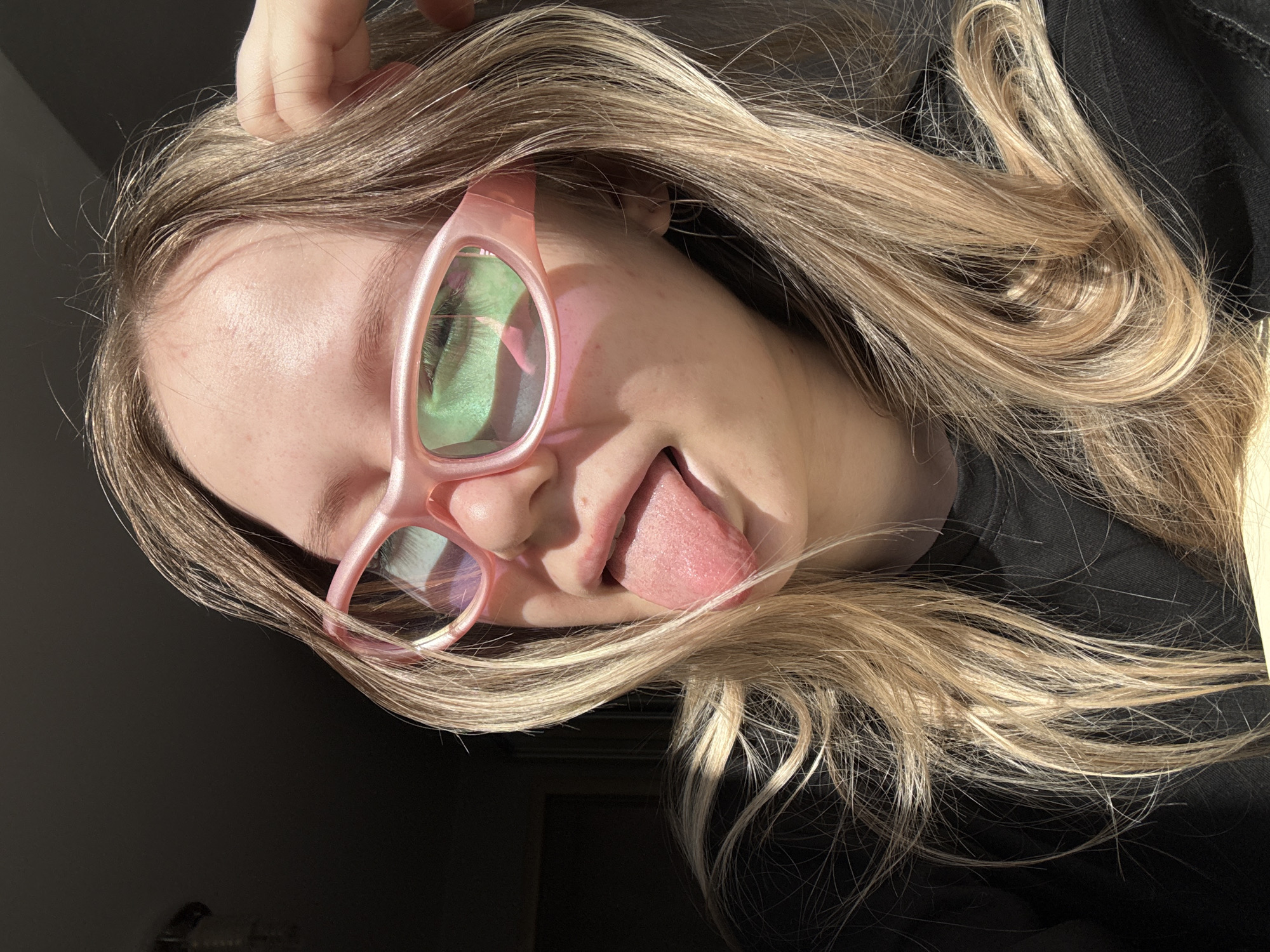 A person with blonde hair, wearing pink-rimmed glasses, is sticking their tongue out. The location is not Zhigalki, Russia. The person is wearing a black shirt. The background is a dark, undefined space. The lighting is bright, casting shadows.