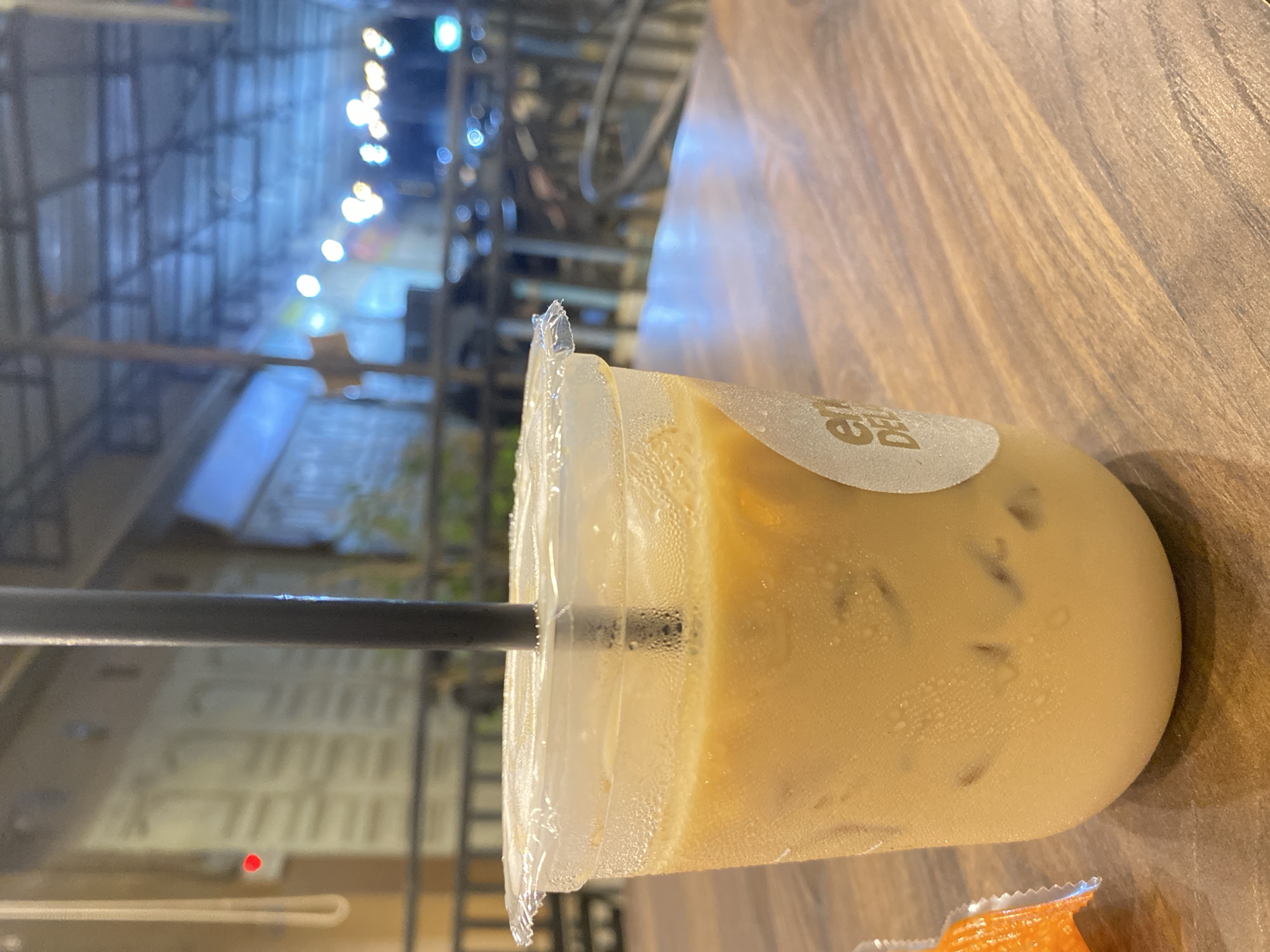 A close-up shot features a clear plastic cup filled with an iced coffee drink, set on a wooden surface. A black straw is inserted into the cup. The cup has a sticker with "EN DELI" visible on it. The background shows an outdoor area with construction scaffolding and streetlights. The blurry background suggests an urban environment. The location is Tanjongkapai, Indonesia. An orange package is partially visible near the bottom edge of the frame.