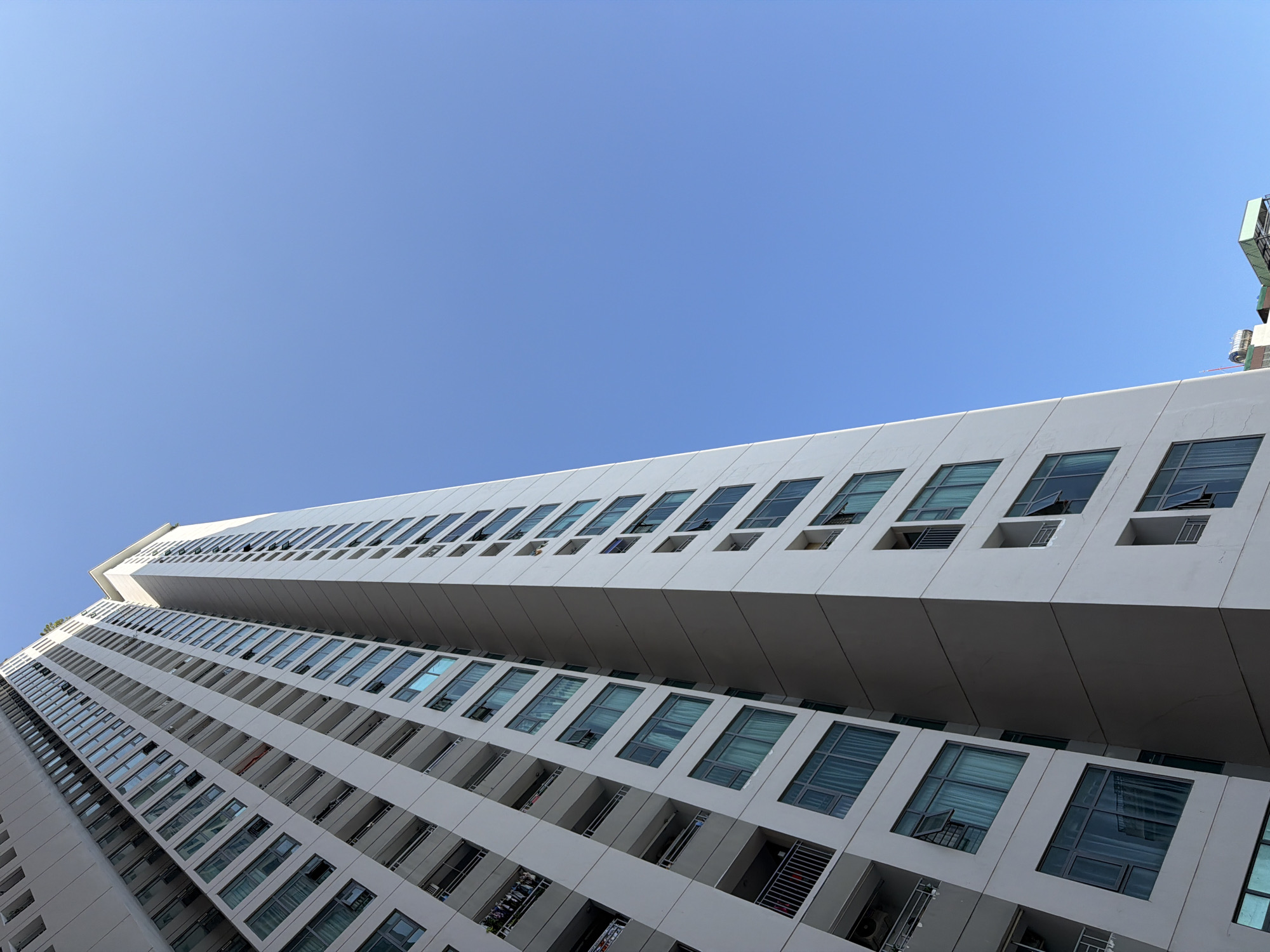 A striking low-angle photograph captures a tall, modern building against a clear, vibrant blue sky. The building, likely an apartment or residential complex, dominates the lower and middle sections of the frame, tapering upwards and seemingly curving or twisting away from the viewer. Its architecture features a clean, white or light grey facade with a distinctive pattern of recessed windows and balconies.
Rows of rectangular windows, some appearing as balconies with railings and others as glass panes, are systematically arranged across the building's multiple stories. Many of the windows and balcony openings are empty, while some show glimpses of interiors or small objects, such as plants on a balcony. The glass in the windows has a reflective, greenish-blue tint, mirroring parts of the sky and light.
The perspective from below emphasizes the building's height and angular design, creating strong diagonal lines that converge towards the top. The sky is uniformly clear and bright blue, suggesting a sunny day. A small, indistinct structure, possibly part of an antenna or rooftop equipment, is visible at the very top right edge of the frame. There are no people or specific activities depicted, and no visible text. The setting is consistent with urban architecture in Bac Nha Trang Ward, Vietnam.