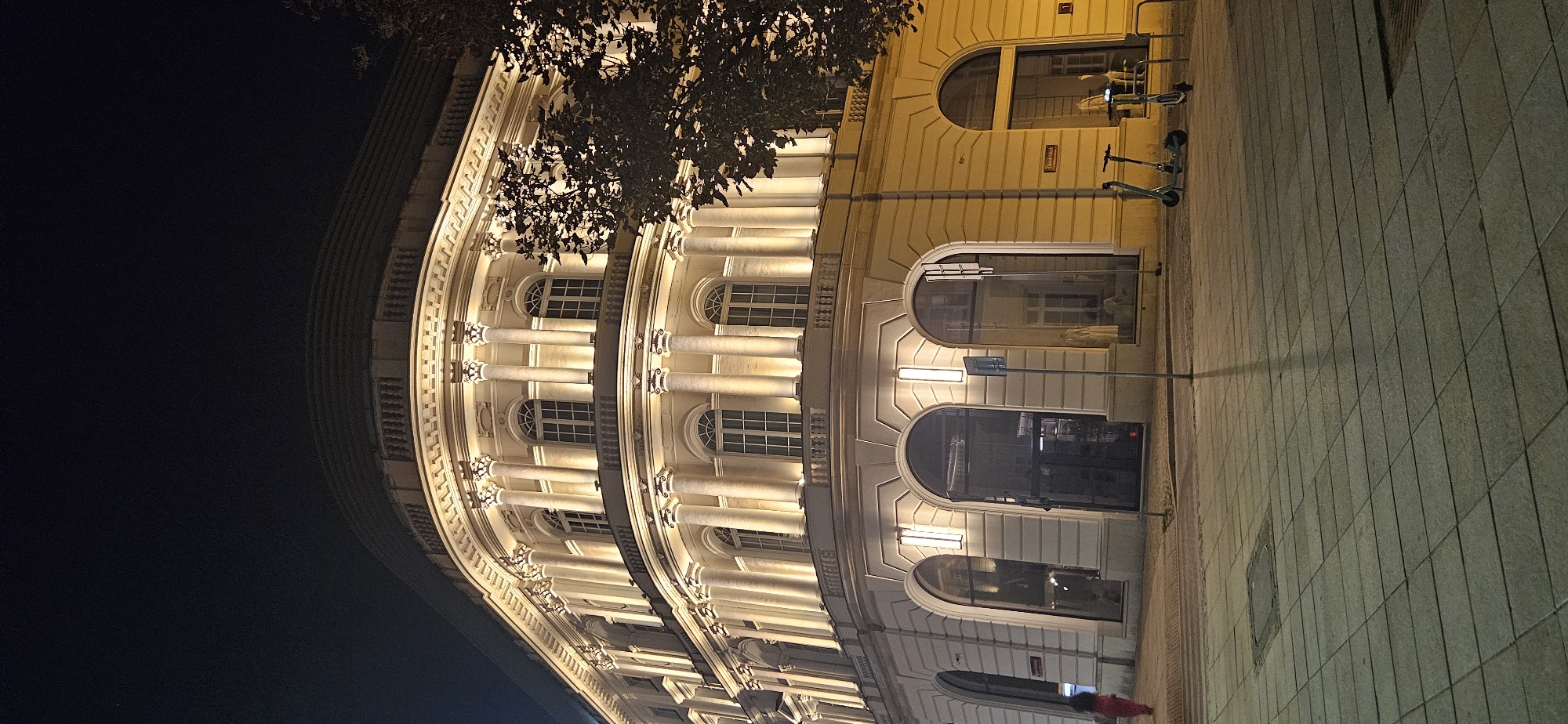 The image captures a nighttime view of a curved, cream-colored building with classical architectural elements, illuminated by warm yellow lights. The building features multiple arched windows on the ground floor and rows of arched windows on the upper floors, each framed by columns. A balustrade runs along the balconies of the upper floors. The sidewalk in front of the building is paved with light-colored square tiles. To the right of the building, on the sidewalk, are two electric scooters parked next to a yellow railing. A street sign with Polish text, possibly "Nowogrodzka," is visible on the building. In the foreground, a portion of the paved sidewalk is shown. A person in a red outfit is walking away from the camera on the left side of the sidewalk. The sky above is dark. The building is located at Nowogrodzka 25 in Warsaw, Poland.