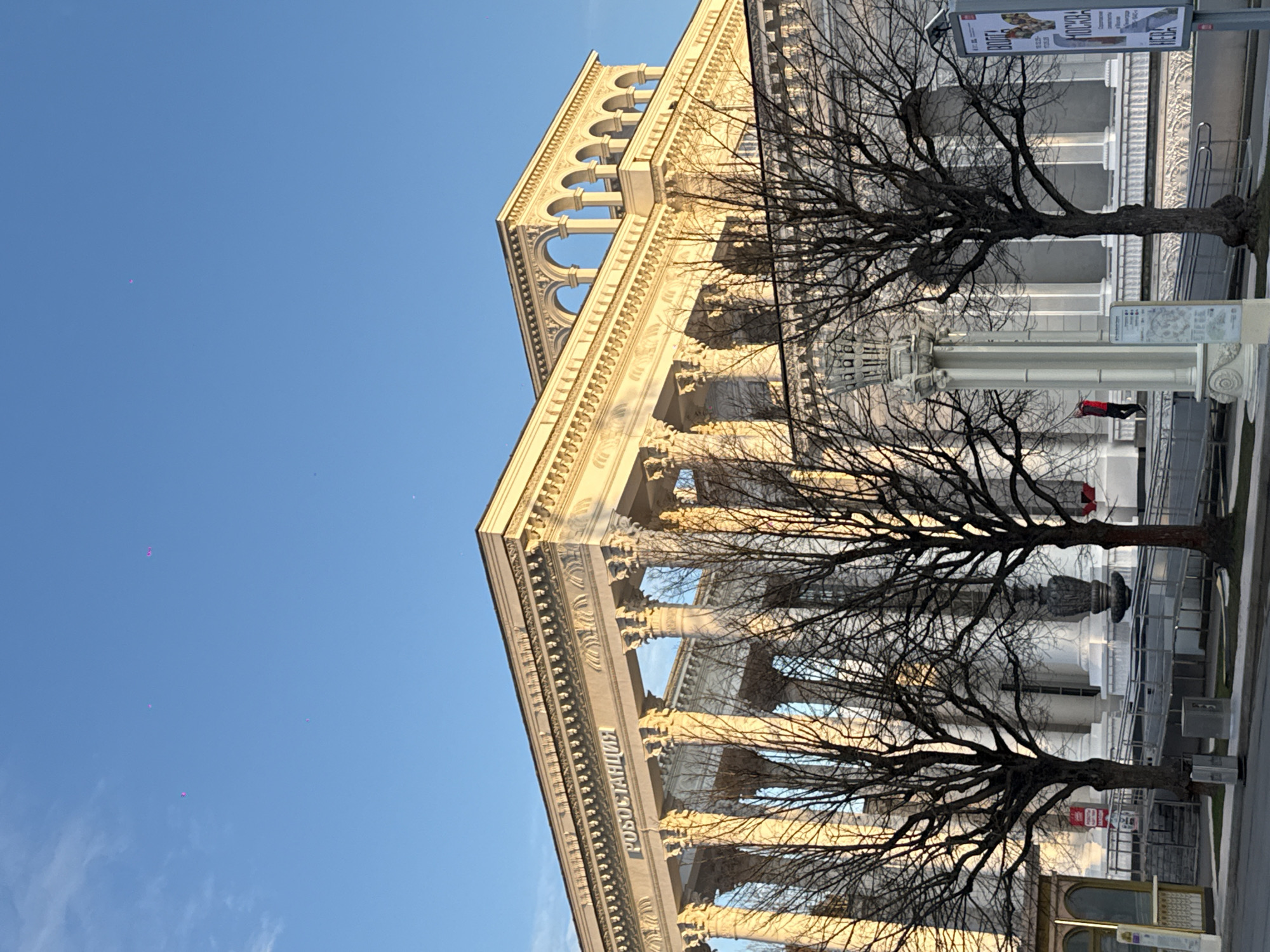 The image shows a building with neoclassical architecture, featuring columns and a balustrade with arches. The building has the word "РОБОСТАНЦИЯ" written on it in Cyrillic. Two bare trees are in the foreground, partially obscuring the building. A person wearing a red jacket can be seen in the background. A street with a bus stop is visible. The image is taken in Moscow, Russia.
