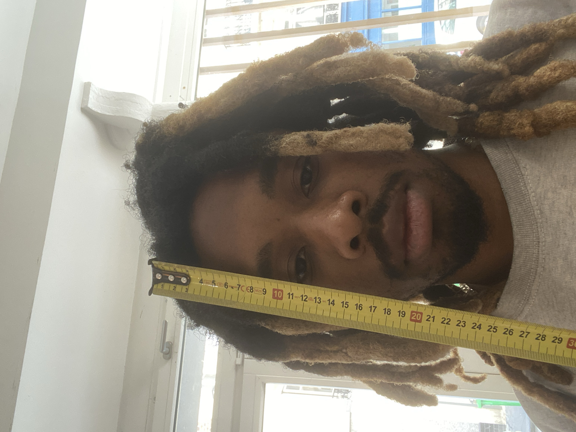 This close-up, vertically oriented image features a person with dark skin and a beard, looking directly at the camera. Their head is tilted to the right, showcasing a substantial volume of thick dreadlocks that transition from dark roots to lighter, possibly dyed, ends. A yellow retractable measuring tape is held horizontally across the person's forehead, resting on their hair. The metal tab of the tape is visible on the far left, near the "1" mark, indicating the starting point of the measurement for the dreadlocks.
The tape displays clear numerical markings in centimeters, visible up to approximately 29 cm, with the numbers "10" and "20" highlighted in red. The person is wearing a grey top, visible on their right side. In the background, a bright window with horizontal blinds or bars lets in natural daylight, illuminating the scene. A white wall or window frame structure is also visible. The setting appears to be an indoor space, contextually located in Paris, France. The person's expression is neutral and direct. The central activity depicted is the precise measurement of the person's hair length.