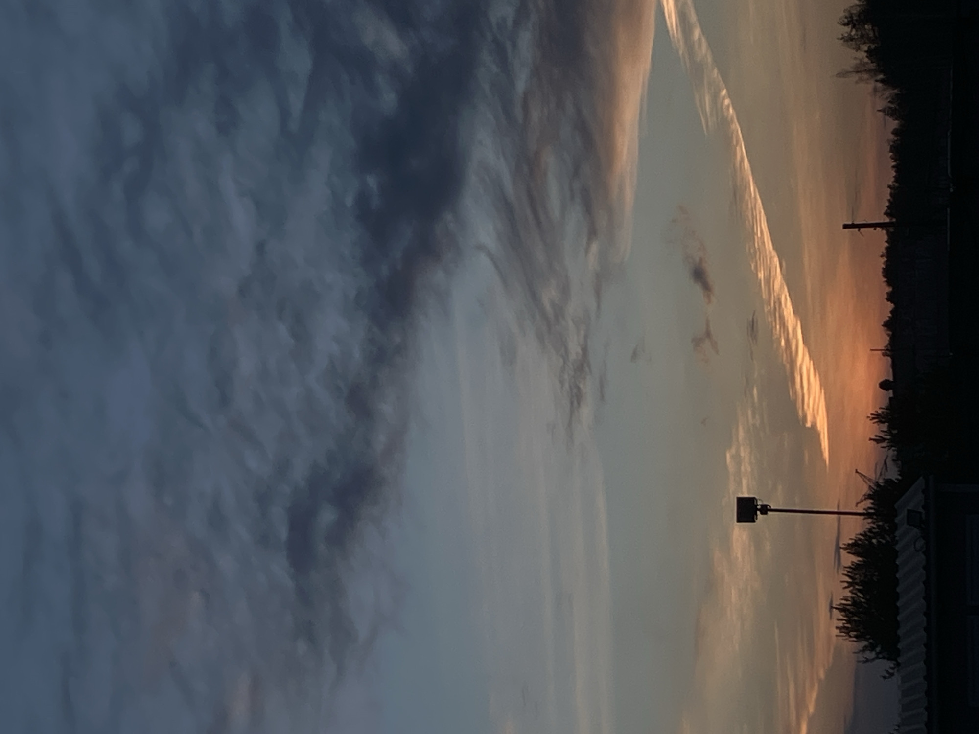 The image shows a sunset sky with clouds ranging from dark blue-gray to orange. A vapor trail bisects the orange portion of the sky. Silhouetted trees and some structures are visible at the lower right edge of the image. A lamp post also is apparent. The image is from an unknown location in Noviy, Russia. No people or interactions are apparent in the picture.