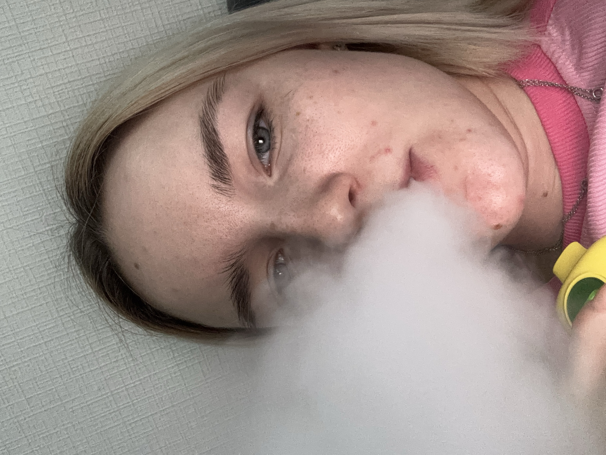 A close-up, eye-level shot presents a young, light-skinned person with blonde hair and blue eyes. They are seen exhaling a large cloud of vapor, obscuring much of their face. The person is wearing a pink sweatshirt and a silver chain necklace is visible around their neck. A yellow and green vaping device is partially visible on the right side of the frame, near their hand. The background is a textured, light grey wall. The location is the city of Unknown, Russia.