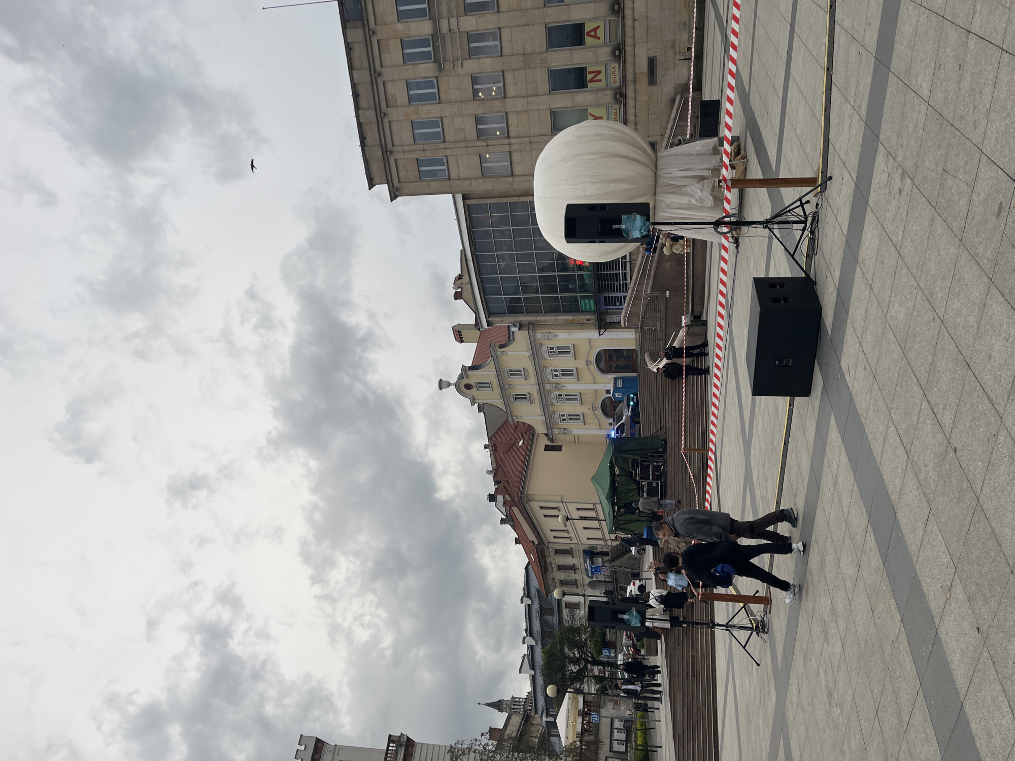 The image captures a scene in a city, possibly Unknown, Unknown, under a cloudy sky. The foreground shows a public square paved with grey stone. A large, white, spherical object, resembling a balloon or light fixture, is placed on a stand. It's flanked by a black speaker. Red and white striped tape marks off an area. Several people walk and stand in the square. A row of people is sitting on benches. In the background are various buildings of different styles and heights. A bird flies in the sky.