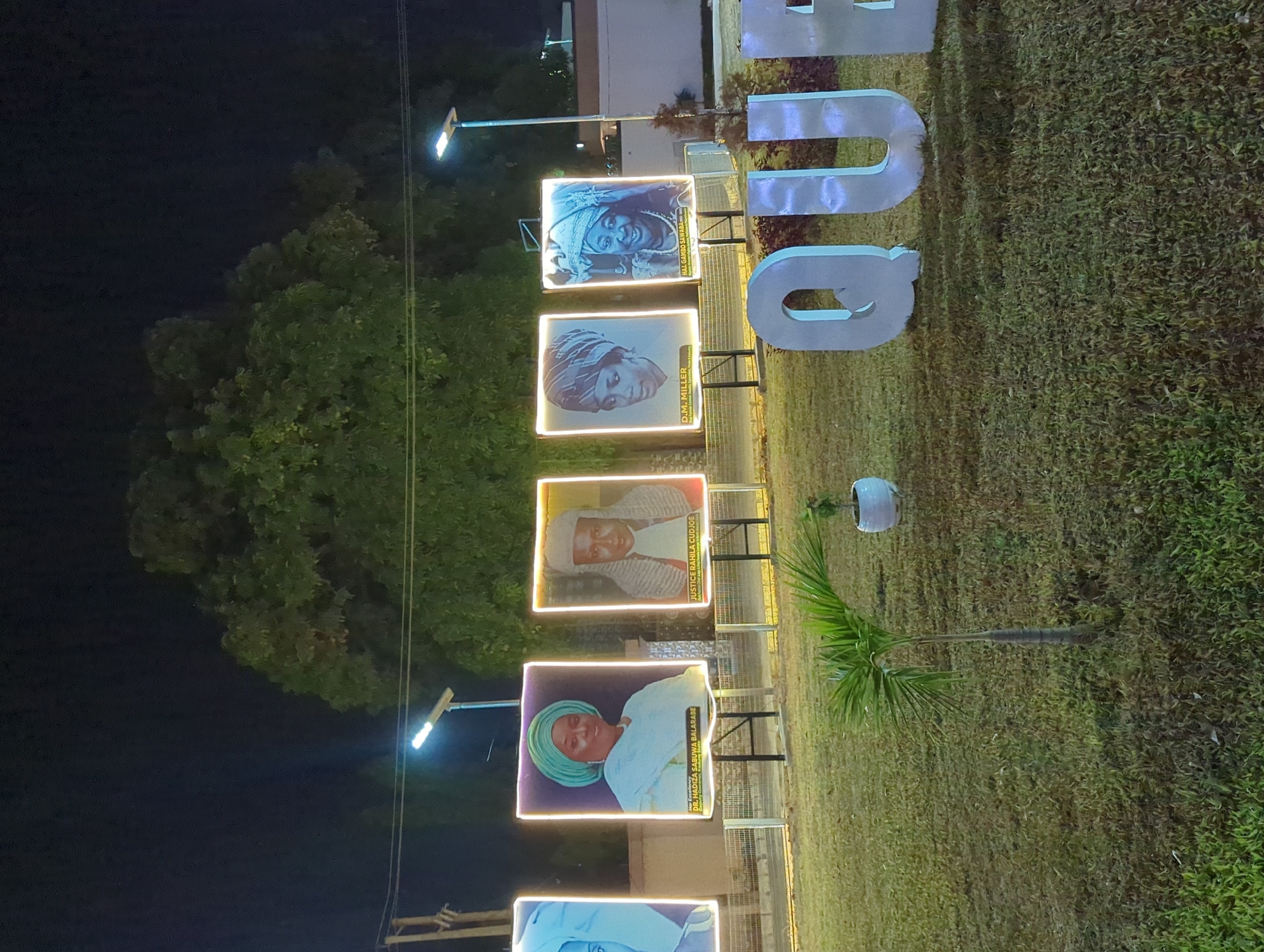 The image shows a night scene in Unguwar Rimi, Nigeria, featuring a vertical display of five illuminated portraits mounted on stands. Each portrait is framed with a light, featuring images of individuals with their names: D.M. Miller, Dr. Hadiza Sabuwa Balarabe, Haj. Gambo Sawaba, Justice Rahila Cudjoe. To the right of the portraits, large block letters, "QU", are visible. Green foliage and a palm tree are also present, suggesting a landscaped area. A street lamp is seen near the portraits, providing light to the scene.