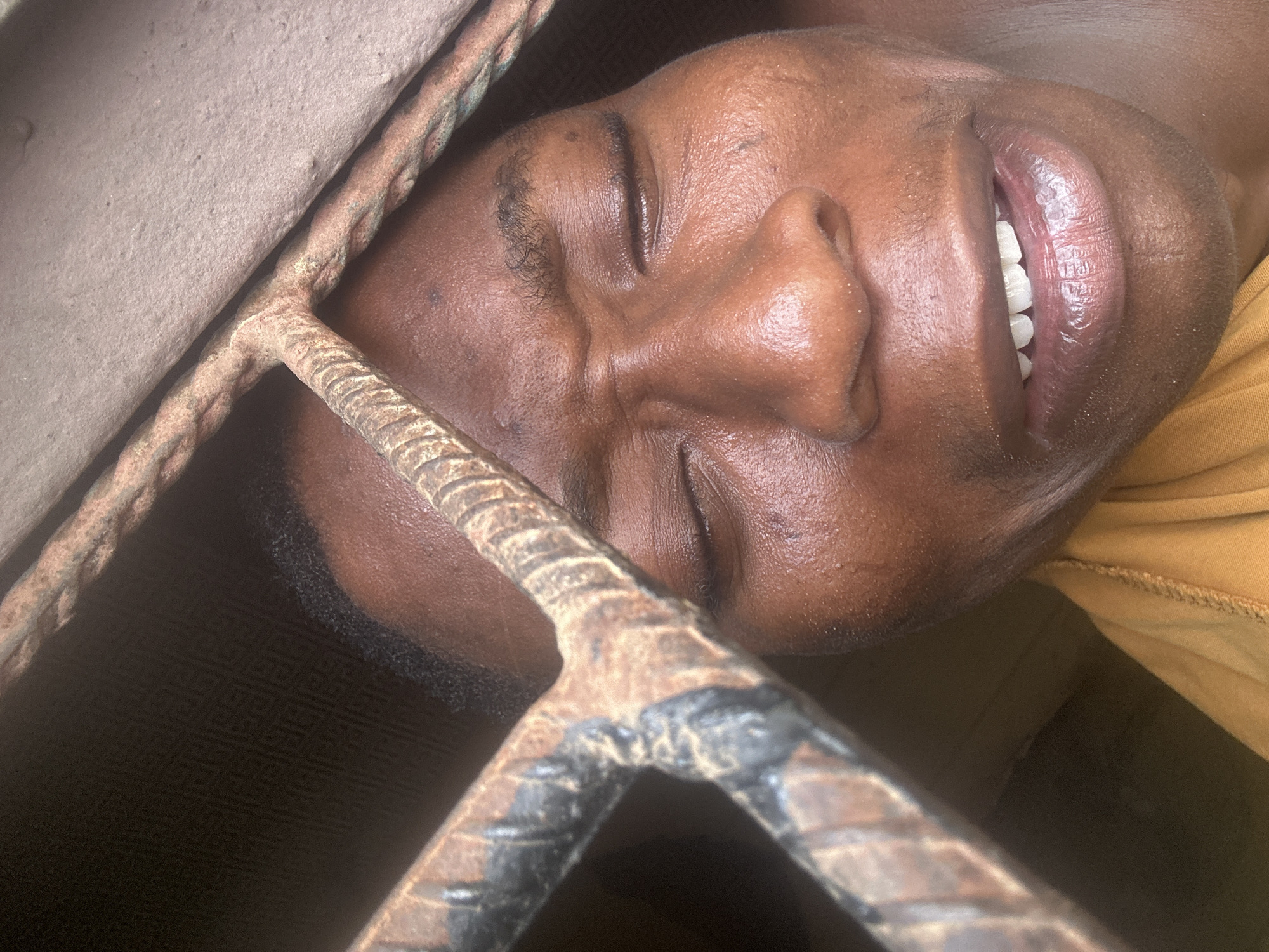 A close-up, high-angle shot captures a person with dark skin, seemingly male, resting or reclining behind a set of rusted metal bars. The person's face is positioned diagonally across the frame, with eyes closed and a soft smile revealing bright white teeth. Their expression suggests peaceful rest or contentment.
Prominent in the foreground are thick, twisted rebar or metal bars, which partially obscure the individual's face. These bars exhibit significant rust and wear. A lighter, rough-textured surface, possibly concrete or stone, is visible in the upper left background, behind the bars. The person's skin appears smooth, and short, dark hair is visible around the temples and jawline. A mustard yellow or light brown garment is visible on their right side. The background directly behind the person is dark and indistinct, hinting at an enclosed or shaded environment. The lighting is soft, highlighting facial features and the textured surfaces of the bars. No text is visible in the image.