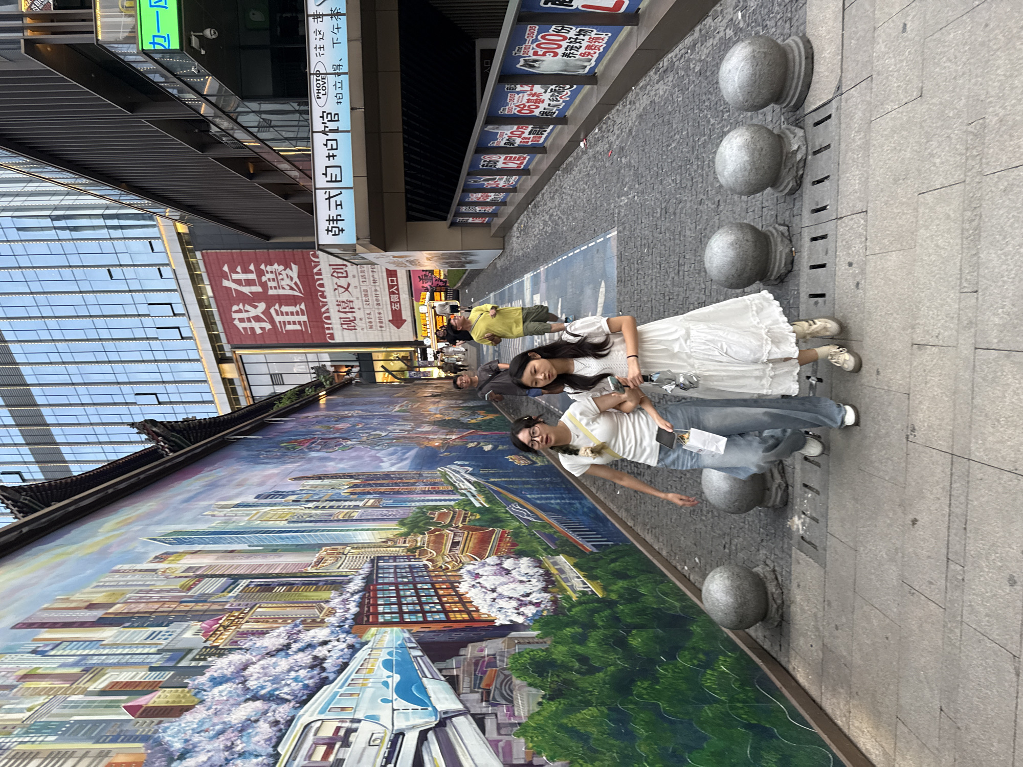 The image depicts a narrow pedestrian walkway in Chongqing, Yuzhong District, China, flanked by a vibrant, detailed mural on the left and modern buildings on the right. Two young women are walking towards the viewer on the paved walkway. The woman on the left wears glasses, a white t-shirt, and blue jeans. She is holding a small paper bag in her left hand. The woman on the right wears a white ruffled dress and is holding a silver umbrella.
The mural on the left wall is a colorful depiction of a city skyline with skyscrapers, traditional Chinese architecture, a monorail, and blooming trees, possibly cherry blossoms. The walkway is paved with cobblestones and has a blue lane marking. On the right side, a multi-story building features glass windows and balconies, with signs in Chinese characters visible. Several spherical stone bollards line the edge of the walkway. The lighting suggests it is late afternoon or early evening. Other pedestrians are visible further down the walkway.