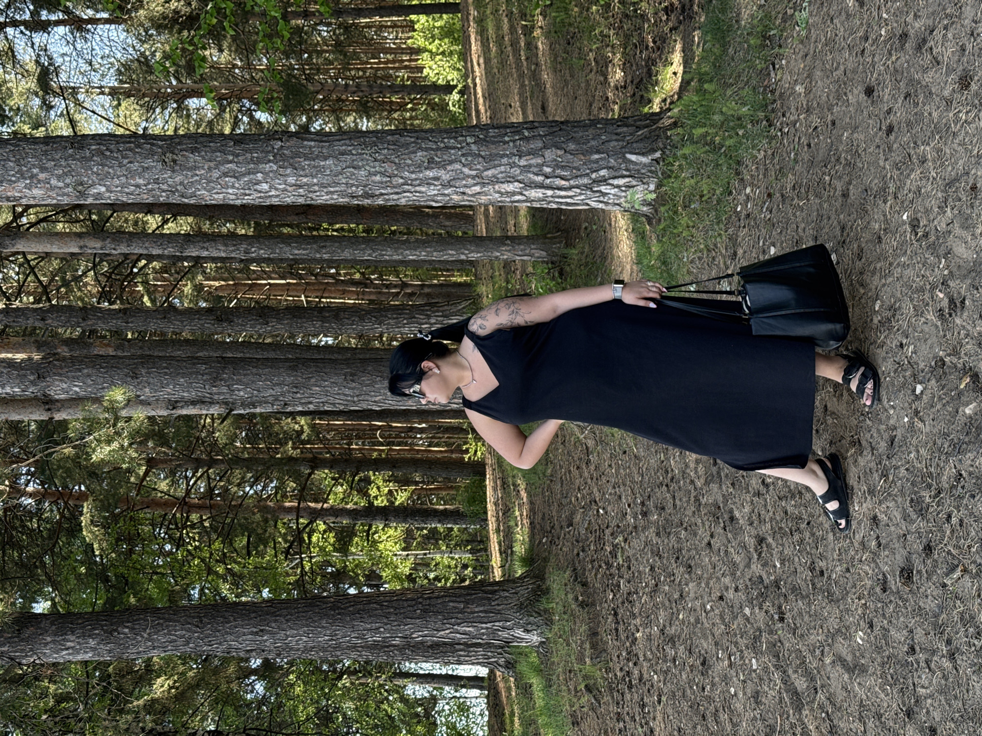 A woman stands on a dirt path in a pine forest. She is wearing a long black dress and black sandals, and she carries a black bag on her shoulder. The location is Chaikovsky, Russia. Pine trees form a dense background.