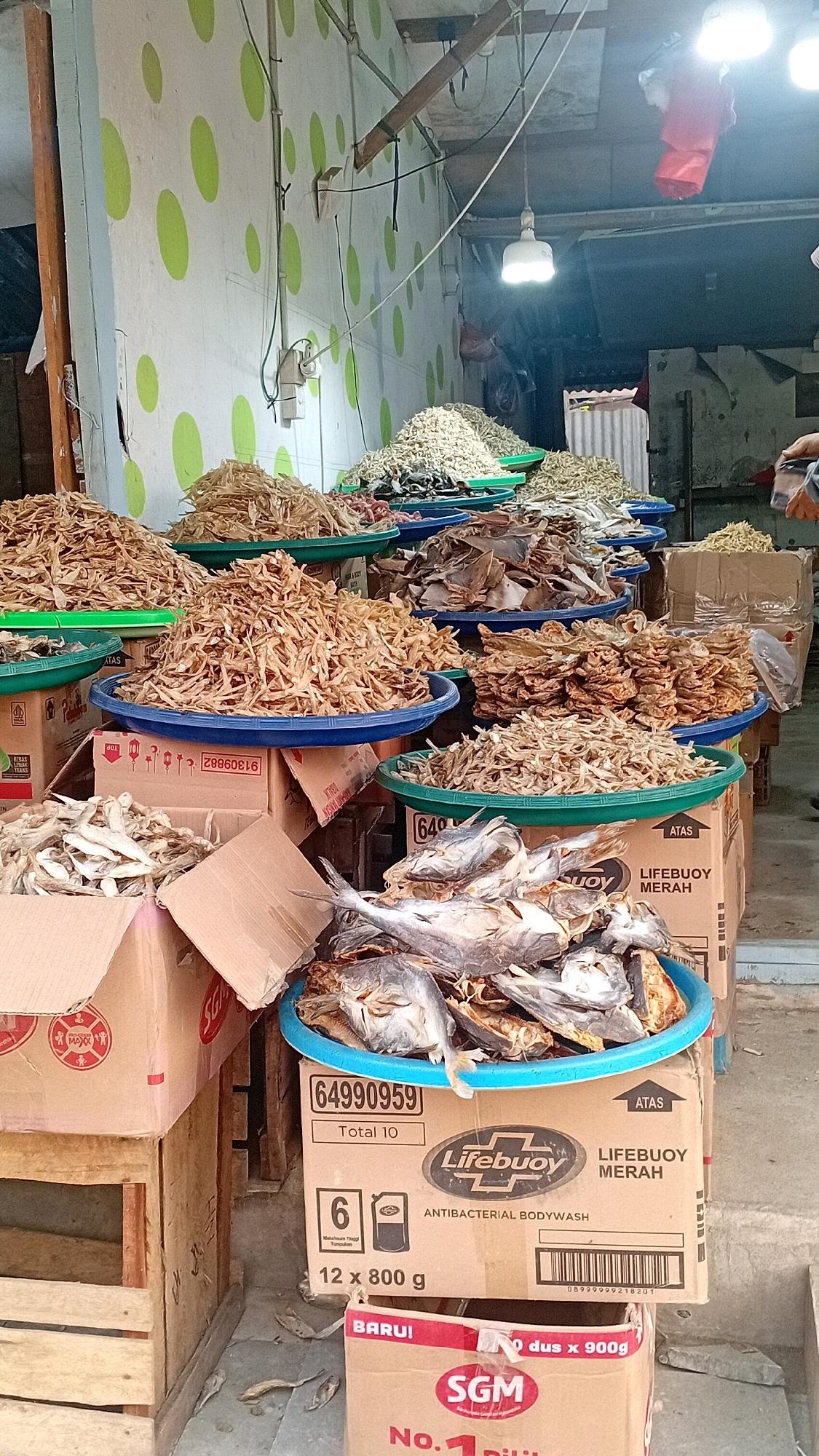 The image depicts an outdoor market stall in Jambi, Indonesia, likely in a traditional market setting. The stall is laden with various dried fish products, arranged in large, shallow green and blue plastic trays. The products include small dried anchovies and larger, flatter dried fish. Several cardboard boxes are stacked beneath the trays, some displaying branding such as "Lifebuoy Merah" and "SGM." A few whole dried fish are visible in a blue tray in the foreground. The stall is situated against a wall painted white with a green polka dot pattern. Electrical wires and lighting fixtures are visible overhead. The ground is paved with concrete. No people are clearly visible in the scene.