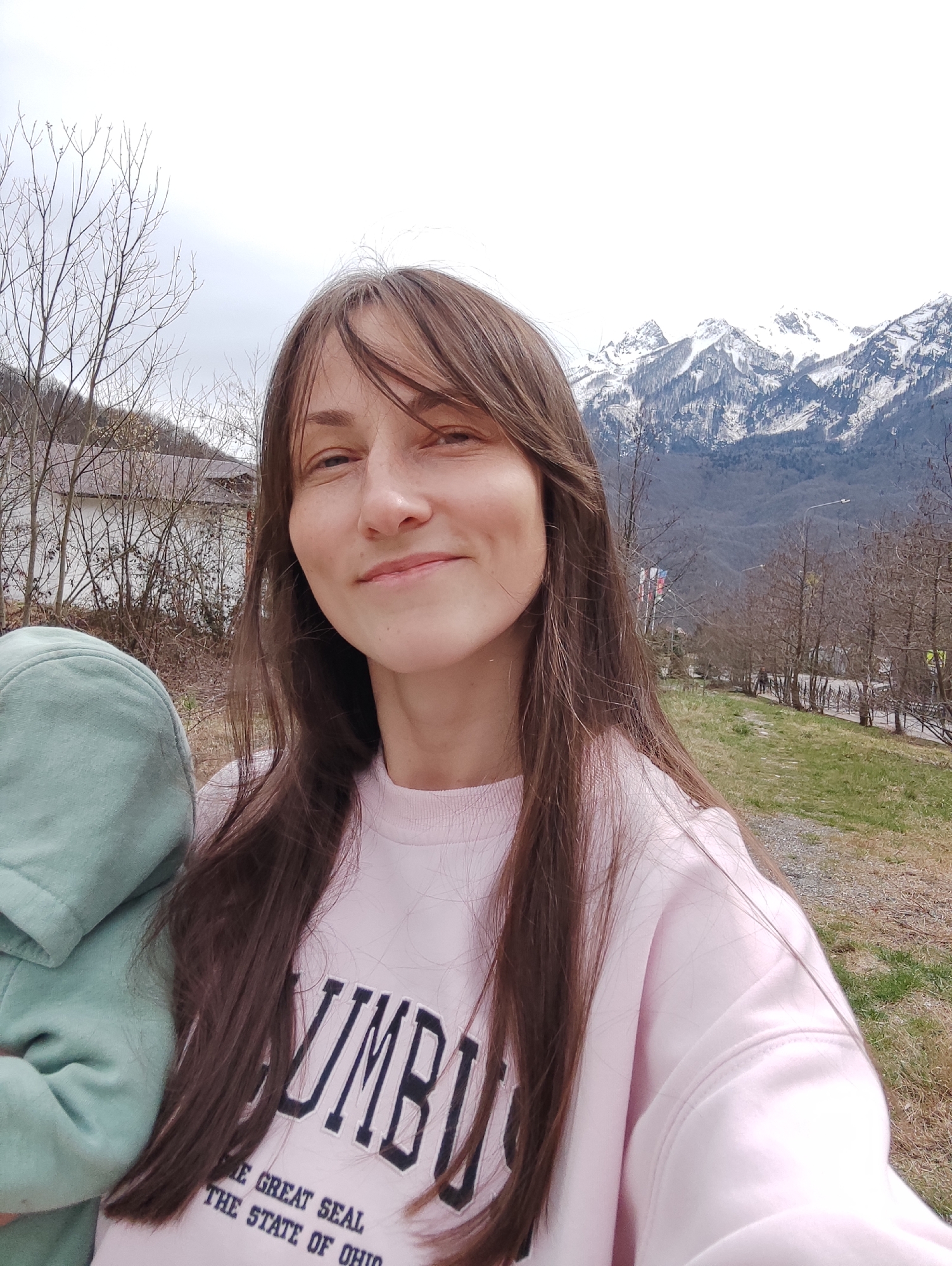 A medium-shot selfie photograph depicts a woman with long brown hair, wearing a pink "COLUMBUS" sweatshirt, standing outdoors. To her left is a child with a green hoodie. Behind her is a landscape of snow-capped mountains and bare trees, with glimpses of buildings and a pathway. Based on the landscape features, this location appears to be in Krasnaya Polyana, Russia.