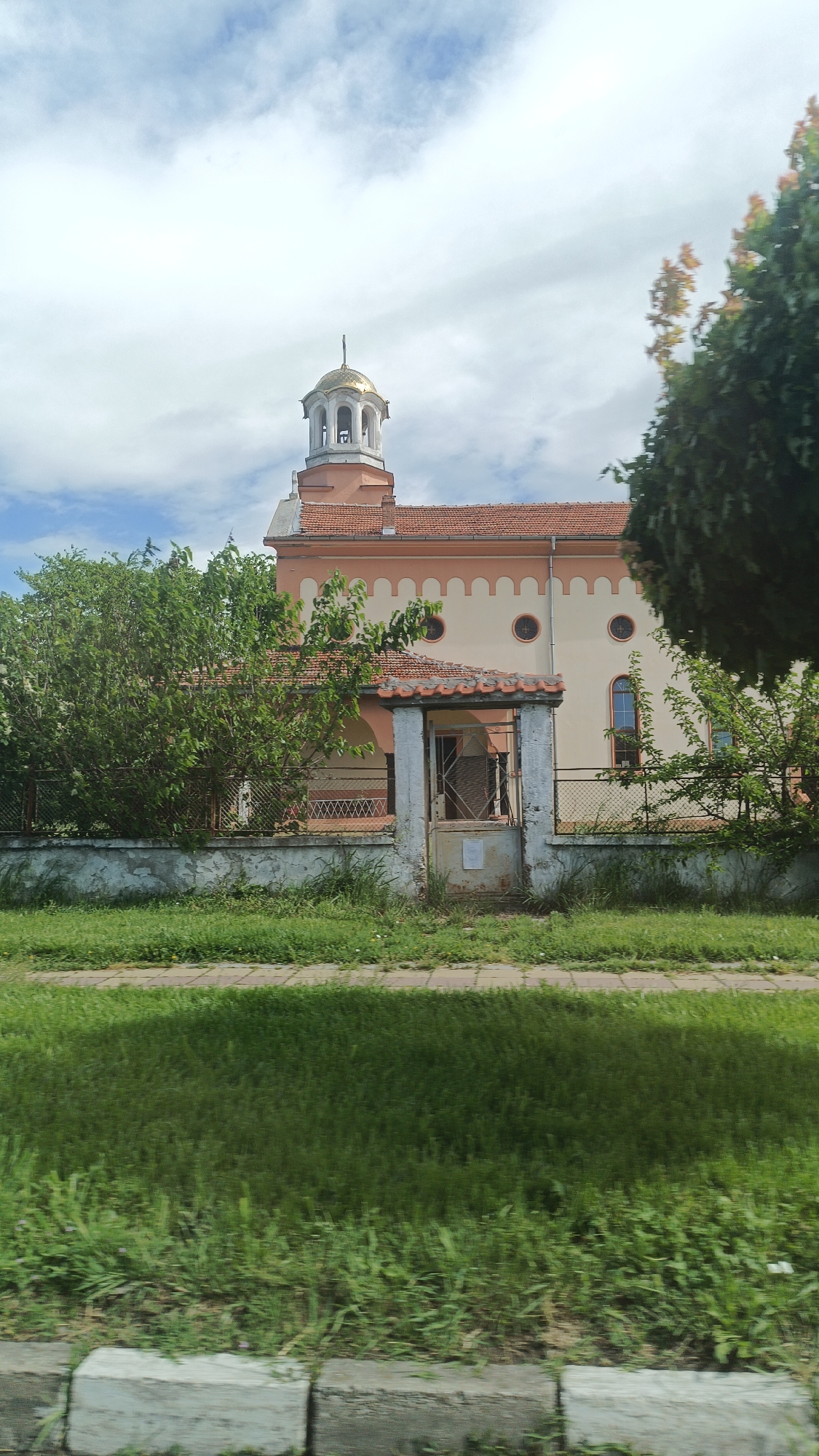 The image captures a view of the Church of Saint Athanasius in the city of Popovitsa, Bulgaria. The church is a one-story building with a golden dome on top. The exterior is painted in light colors with rounded patterns just below the red roof. The church is surrounded by green trees and shrubs, partially obscuring the view of the structure and the weathered fence around it. An open gateway with a tiled roof stands in front of the church, with a rusty metal gate visible through the opening. In the foreground, there is a grassy area and a sidewalk.