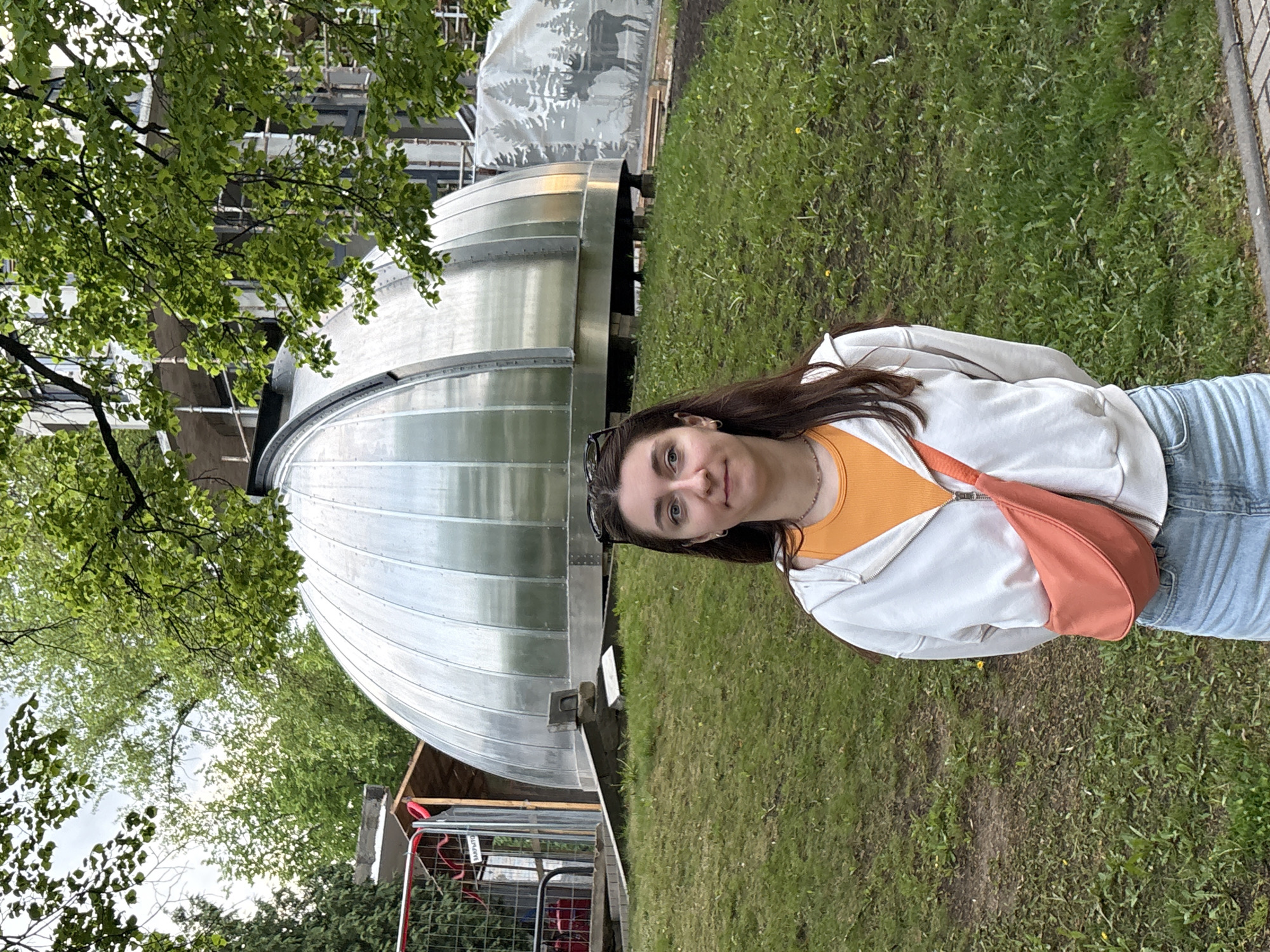 A young woman stands in a grassy area in Minsk, Belarus. A silver, domed structure resembling an observatory is behind her. The structure has a corrugated metal exterior and sits on a metal base. Another similar structure with a moose emblem painted on its side is partially visible behind the first. A metal fence and some greenery are to the lower left of the frame. The woman is wearing a white hoodie over an orange shirt, blue jeans, and an orange shoulder bag. Sunglasses are perched on her head. She is looking directly at the camera. The background also features trees and building elements.