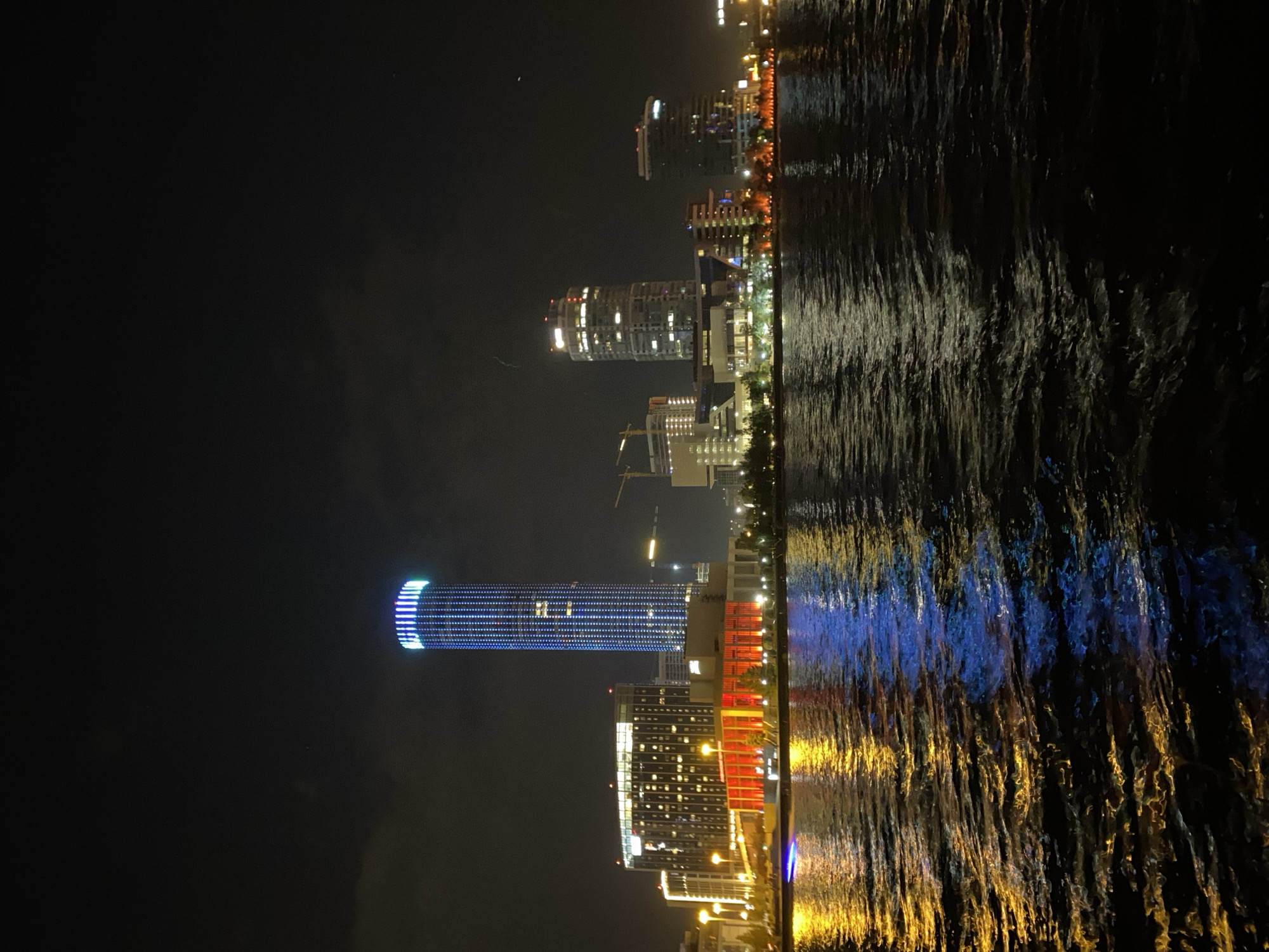 This photograph depicts a nighttime view of the city of Yekaterinburg, Russia, specifically from the Iset River. The scene features a skyline of illuminated buildings reflected in the dark, rippling water. Prominent among the buildings is the Iset Tower, a skyscraper distinguished by its cylindrical shape and blue lighting. Other buildings are also visible, exhibiting a variety of colors, including white, orange, and red. The river's surface reflects the city lights, creating streaks of color. The sky is dark, with some faint lights visible in the distance. A crane is visible near one of the buildings.