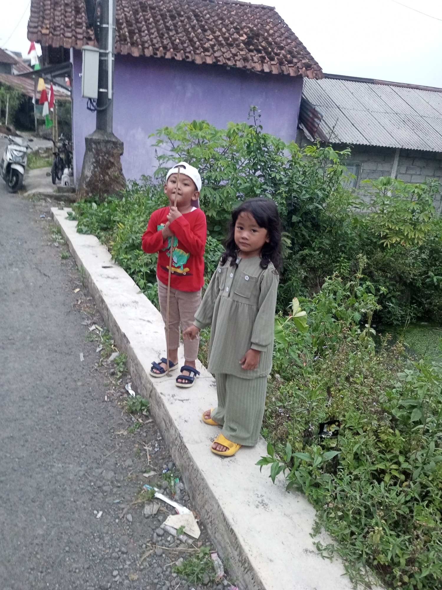 Two young children, a boy and a girl, stand on a concrete ledge next to a paved road. The boy, wearing a red long-sleeved shirt, beige pants, a white baseball cap, and dark sandals, holds a thin stick. The girl, in a light green matching set of shirt and pants and yellow sandals, stands to his right. Behind them, a purple building with a brown tiled roof is visible, along with lush green foliage and a grey corrugated metal roof on another structure. A white motorcycle is parked on the left side of the road. The scene appears to be an outdoor environment in a residential area.