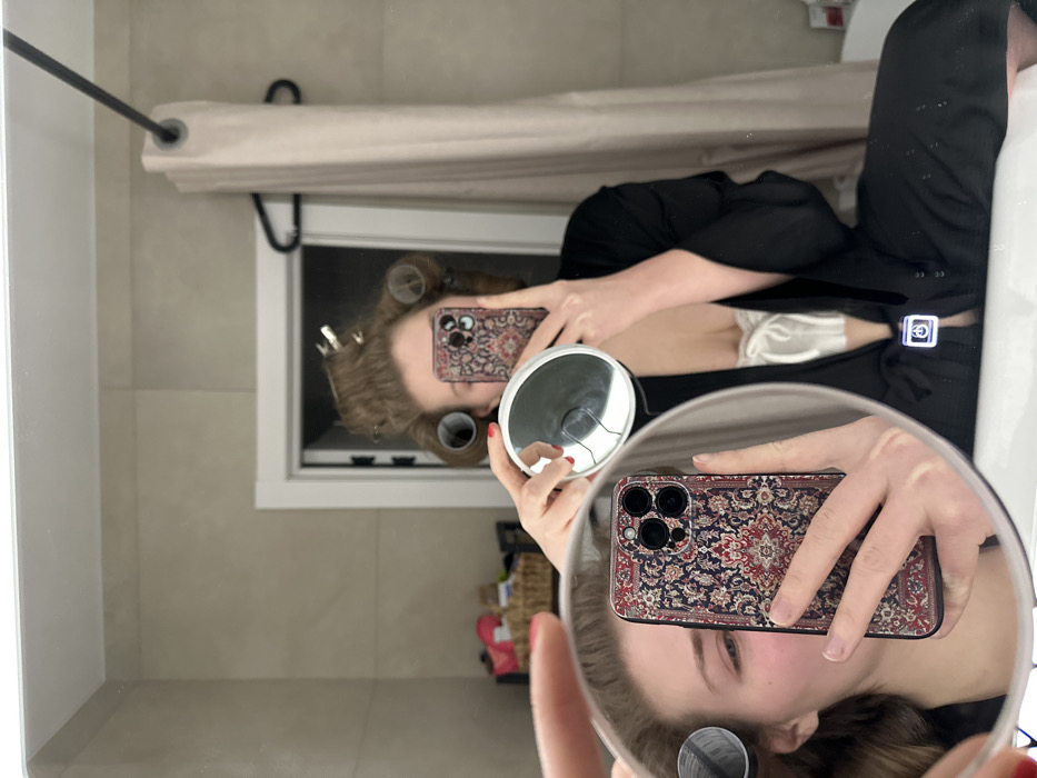 A person in curlers, a black robe, and satin shorts is taking a selfie in a bathroom mirror. They hold a small handheld mirror in one hand and a phone with a patterned case in the other. The phone's reflection is visible in the handheld mirror. A window, shower curtain, and sink are also visible in the background.