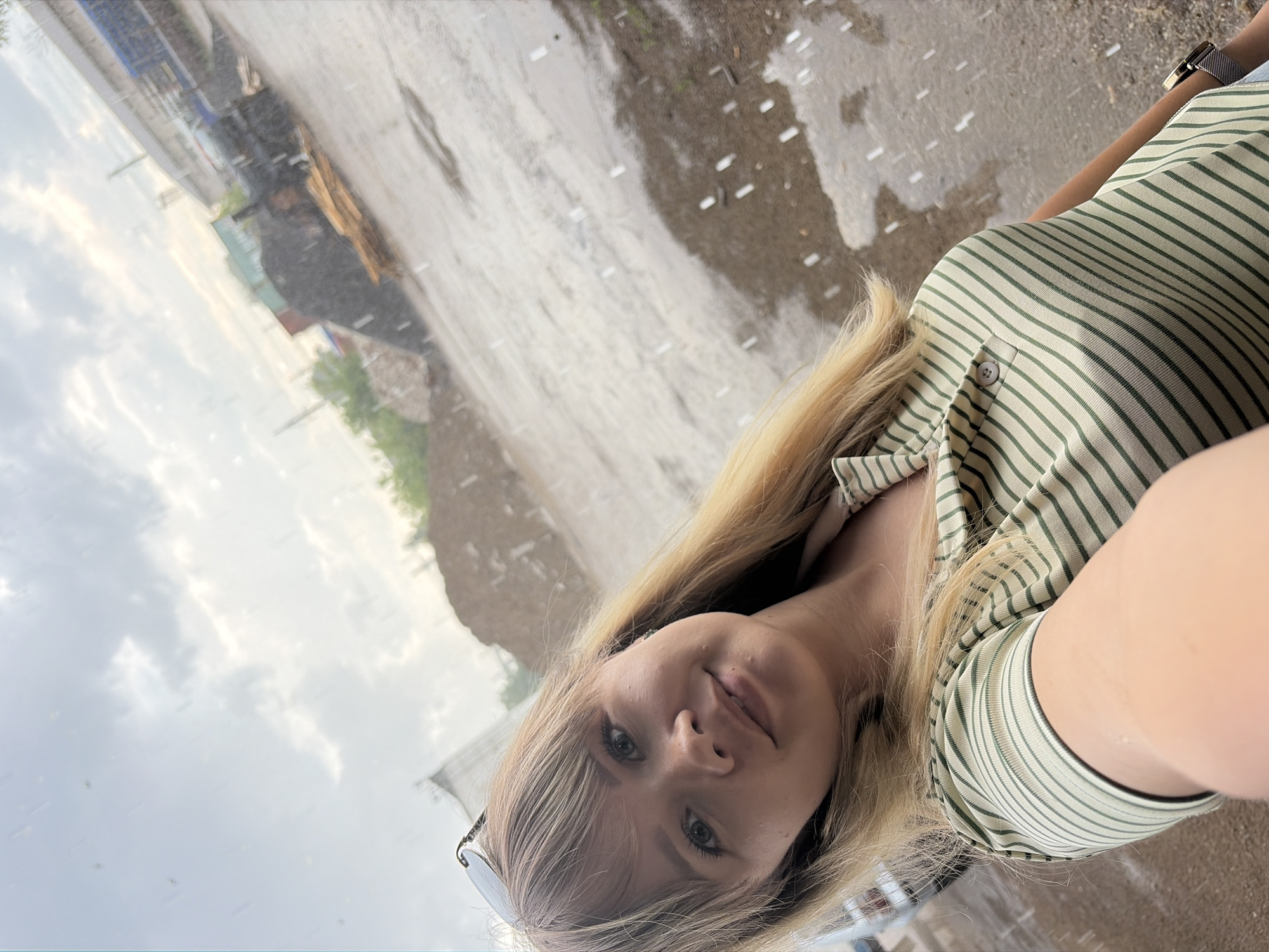 A young woman with blonde hair is taking a selfie in an outdoor setting. She is wearing a striped shirt and sunglasses. The background appears to be a wet, possibly paved area with some industrial structures or debris in the distance. The lighting suggests it might be daytime, with overcast or cloudy skies contributing to a diffused light. There are visible water droplets in the air, indicating it might be raining or has recently rained, making the ground wet and reflective. The overall impression is casual and candid.
