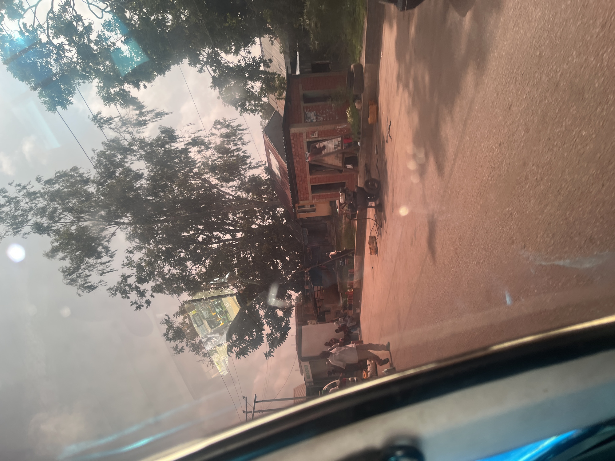 The image depicts a street scene in the city of Bukuru, Nigeria. A paved road runs through the frame, bordered by buildings and trees. Several individuals are visible on the roadside, some standing and others seated. A man in a light-colored shirt and pants is walking away from the camera. Adjacent to him, a group of people are gathered near a low wall. Further down the street, a small brick building with a corrugated metal roof appears to be a shop or a stall. In front of this structure, there are various objects including what looks like a generator or air compressor, some fuel canisters, and discarded tires. Trees with dense foliage line the sides of the street, and power lines are visible overhead. The sky is overcast, with sunlight creating lens flares and reflections on what appears to be a vehicle's window, suggesting the photo was taken from inside a car. The overall impression is of a bustling local area with commercial activity.