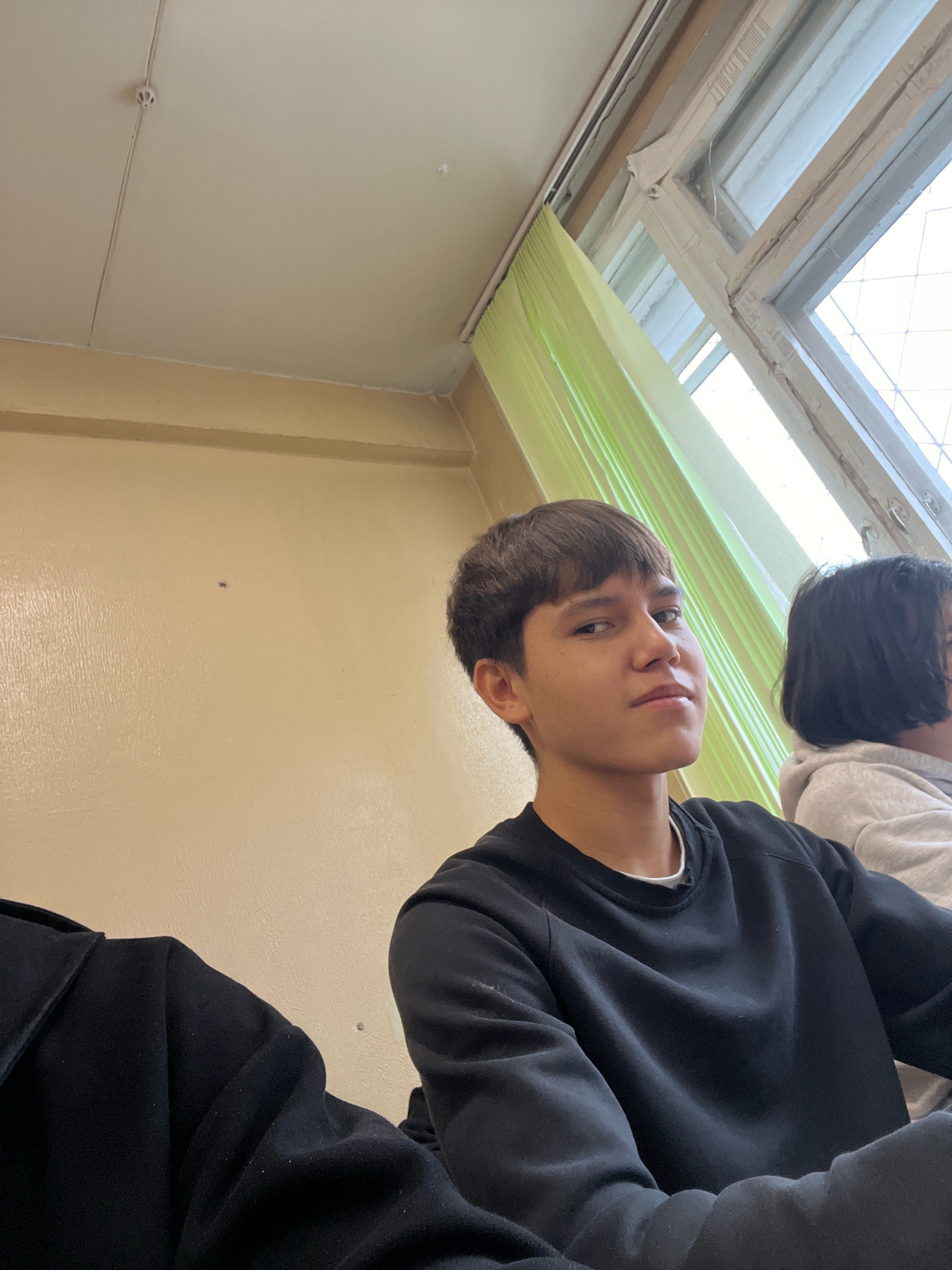 This is a medium-angle, eye-level selfie taken indoors, likely in a classroom in Ufa, Russia. The primary subject is a young male with short, dark brown hair and fair skin, wearing a black crewneck sweatshirt. He is looking towards the camera with a neutral expression. To his right, partially visible, is another person, also appearing to be young, wearing a light-colored hoodie. The background consists of a light yellow wall, a window with a light green curtain, and what appears to be a white ceiling with a fire alarm. The overall environment suggests a casual, everyday setting.