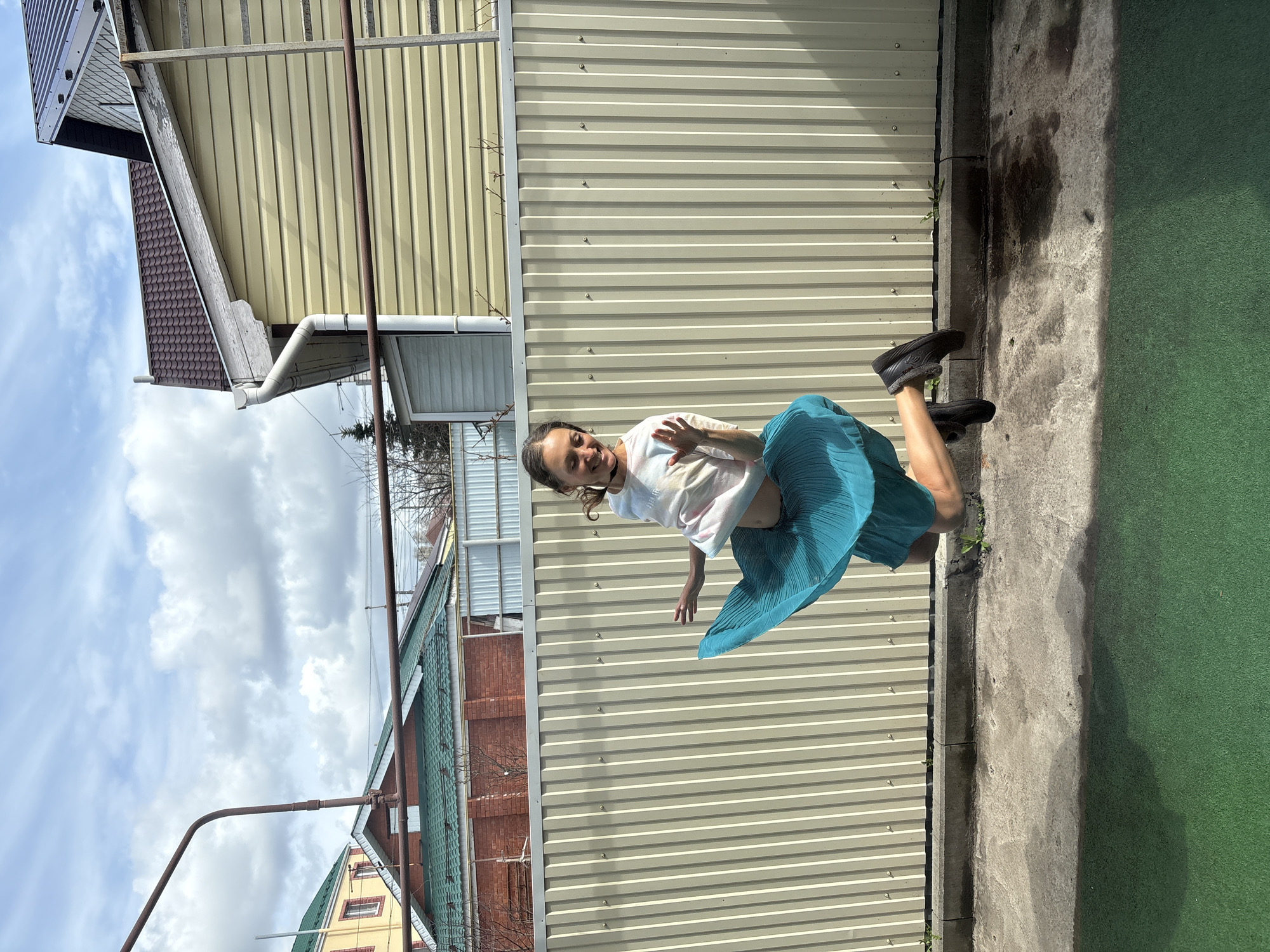 A young woman is standing outside near a metal fence and some buildings. She wears a white shirt and a teal skirt with black shoes. She has her hands up in what appears to be a greeting or gesture. The background shows buildings, some with brick and some with metal siding, under a cloudy sky. The ground has a section of green artificial turf and a concrete strip. I am unable to determine from the image whether this location is in Ulyanovsk, Russia.