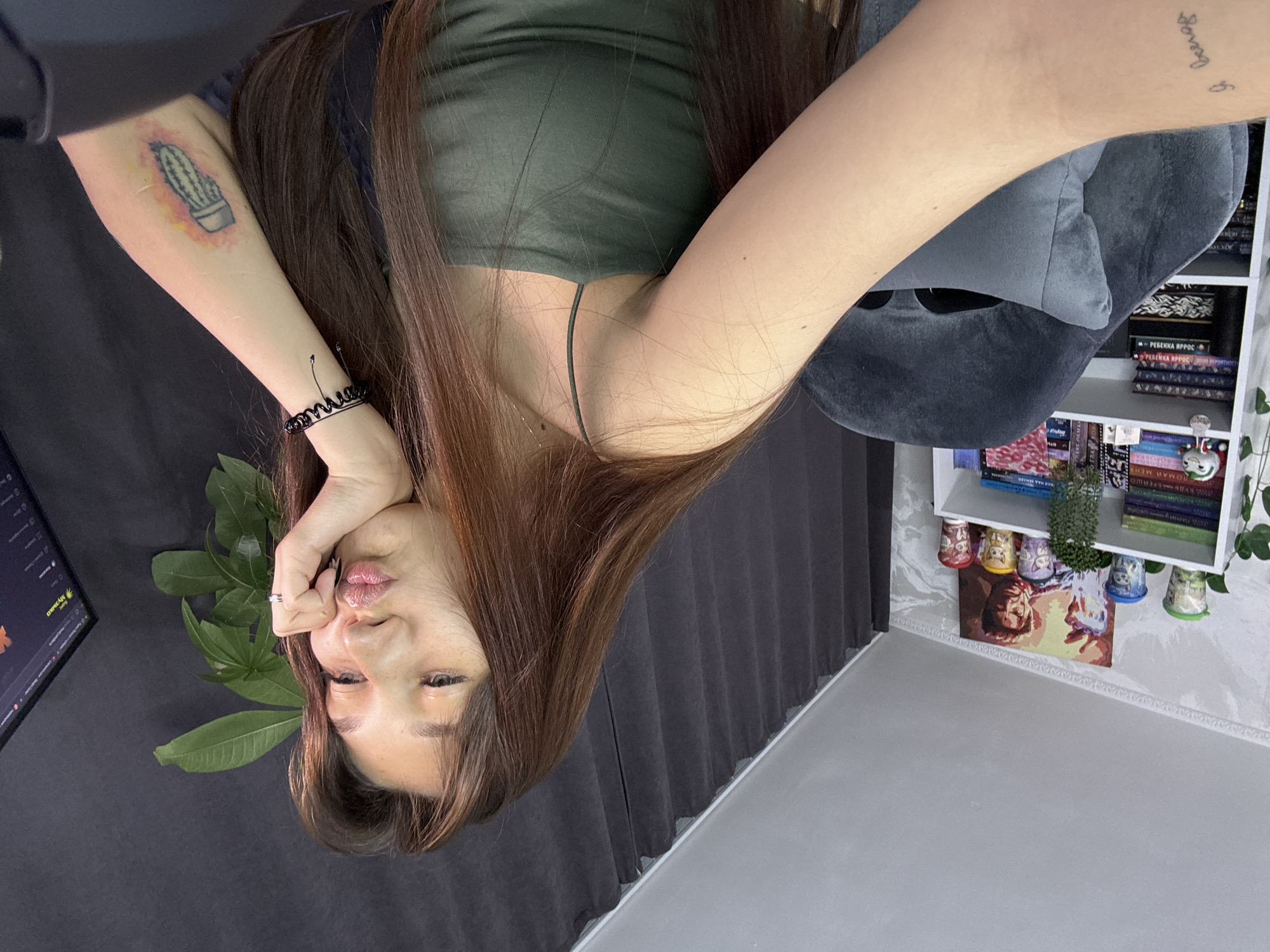 A young woman with long brown hair is sitting in a dark grey chair in front of a computer. She is wearing a green tank top and has a cactus tattoo on her right forearm and a script tattoo on her left forearm. The woman is puckering her lips and resting her chin on her hand. Behind her, a bookshelf is filled with books and decorative cups. A dark grey curtain hangs on the right side of the frame. The scene appears to be indoors, likely a home office or bedroom, and is not identifiable as being specifically in the city of Novokuznetsk, Russia.