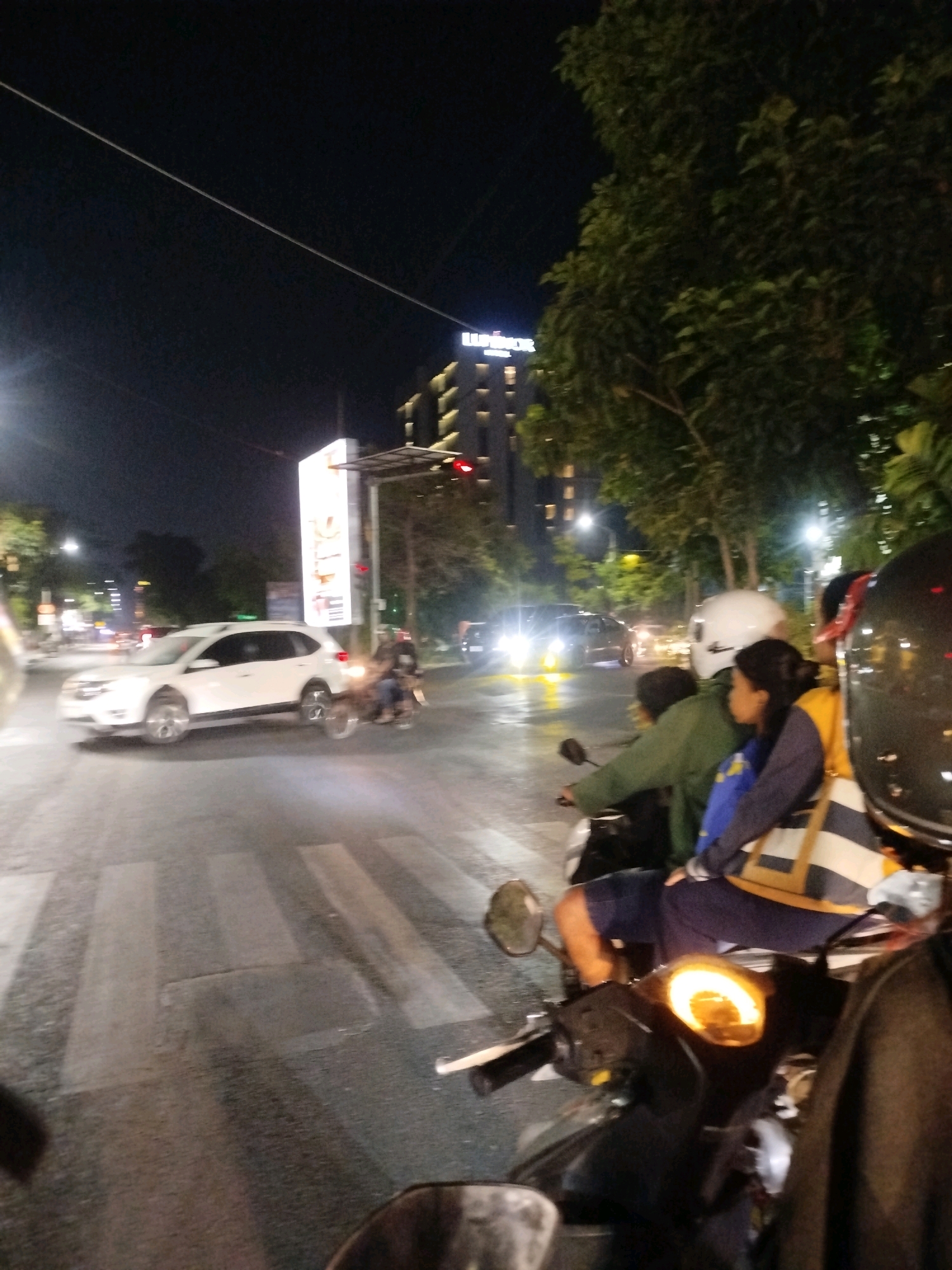 This image captures a nighttime street scene in Surabaya, Indonesia, likely near a busy intersection with a prominent tall building in the background. A white SUV is in motion on the asphalt road, which features a pedestrian crossing. In the foreground, a group of people are on a motorcycle, appearing to be stopped at a traffic light. The individuals are dressed in casual clothing, and one person in the back is wearing a blue jacket. The lighting suggests it's dark, with streetlights and vehicle headlights illuminating the scene. The overall atmosphere is that of urban nightlife, with traffic and people moving about.