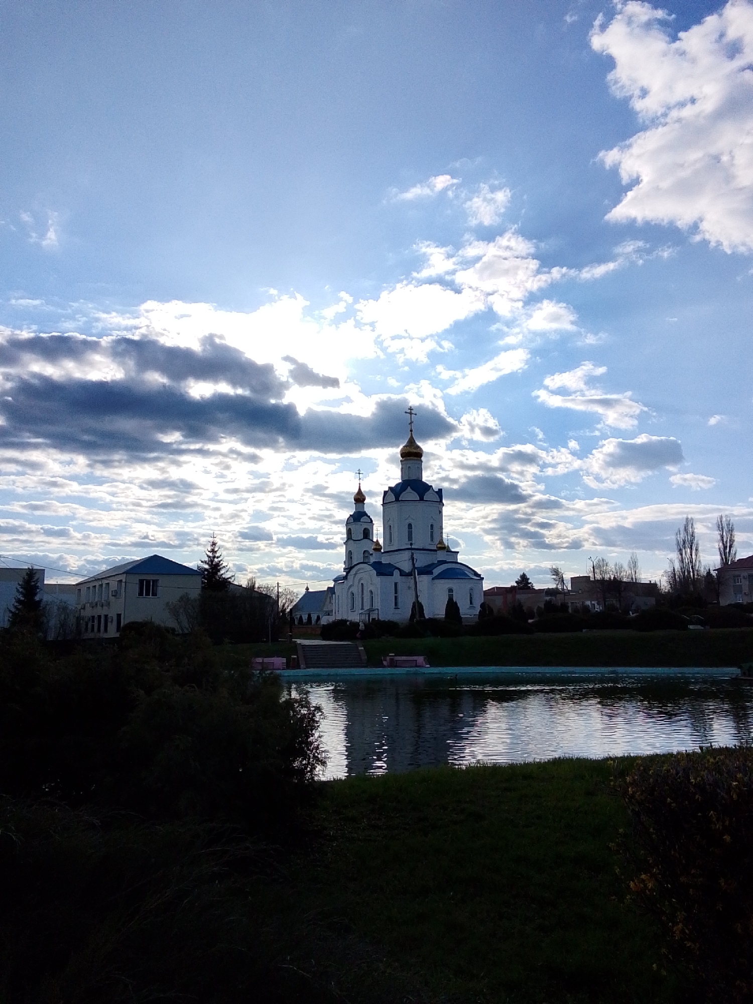The image depicts a white Orthodox church with blue domes and gold crosses, reflected in a pond in Khlevnoye, Russia. The church is situated in a park-like setting, with green grass bordering the pond. There are other buildings visible in the background, including a two-story building with a blue roof. The sky is mostly blue with scattered clouds. The foreground is heavily shadowed.