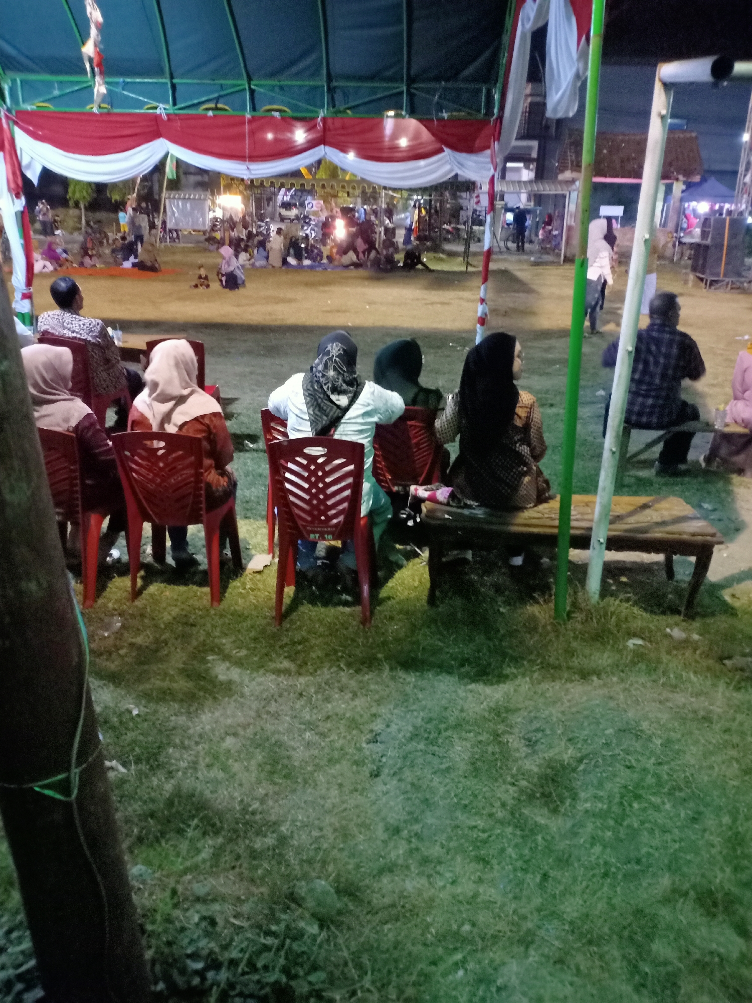 In the city of Sumber Tlaseh, Indonesia, a twilight scene unfolds under a large, blue tent adorned with red and white draped fabric. The foreground is dominated by a grassy area with a rough, brown wooden bench placed on its left side. A dark brown, vertical tree trunk is positioned to the far left of the frame. Several individuals are seated on red plastic chairs facing away from the camera, towards the activity under the tent.
To the left, a person wearing a patterned fabric sits on a chair. Next to them, two individuals wear light brown headscarves, one in a maroon top and the other in a rust-colored top. Further to the right, a person in a light green shirt and a black and white patterned headscarf is seated. Adjacent to them, a person wearing a black headscarf and a patterned outfit sits on the wooden bench. Another individual in a dark plaid shirt is also seated on the bench to the far right, facing away from the camera. Beyond the seated individuals, a crowd is gathered under the tent, with lights illuminating the area and suggesting an event or gathering is taking place. Some people are standing and interacting, while others are seated or observing. Buildings are visible in the background, some with illuminated windows. The overall atmosphere appears to be one of a community event or celebration held outdoors.