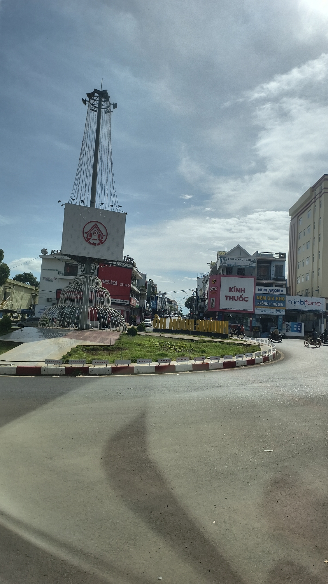 The image captures a street scene in Buon Ho, Vietnam, featuring a roundabout in the foreground. Dominating the scene is a tall, slender tower topped with a large square billboard displaying a red and white logo and text. The billboard is suspended by cables. Below the billboard is an ornamental, tiered metal structure resembling a fountain or abstract sculpture.
The roundabout has a red and white striped curb. Beyond the roundabout, buildings line the street. One prominent building has a "Viettel store" sign. Another building displays "Kính Thuốc" (pharmacy). Further down the street, a large, light-colored building with multiple windows is visible. A sign reading "mobifone" is also present.
In the center of the roundabout, a grass area displays yellow lettering that reads "THÀNH PHỐ BUÔN HÔ" (City of Buon Ho). Several motorcycles are visible on the road, with two riders on one motorcycle and another single rider on a separate motorcycle. The sky is partly cloudy. Shadows of structures, likely from the surrounding buildings and the central sculpture, are cast on the asphalt road.