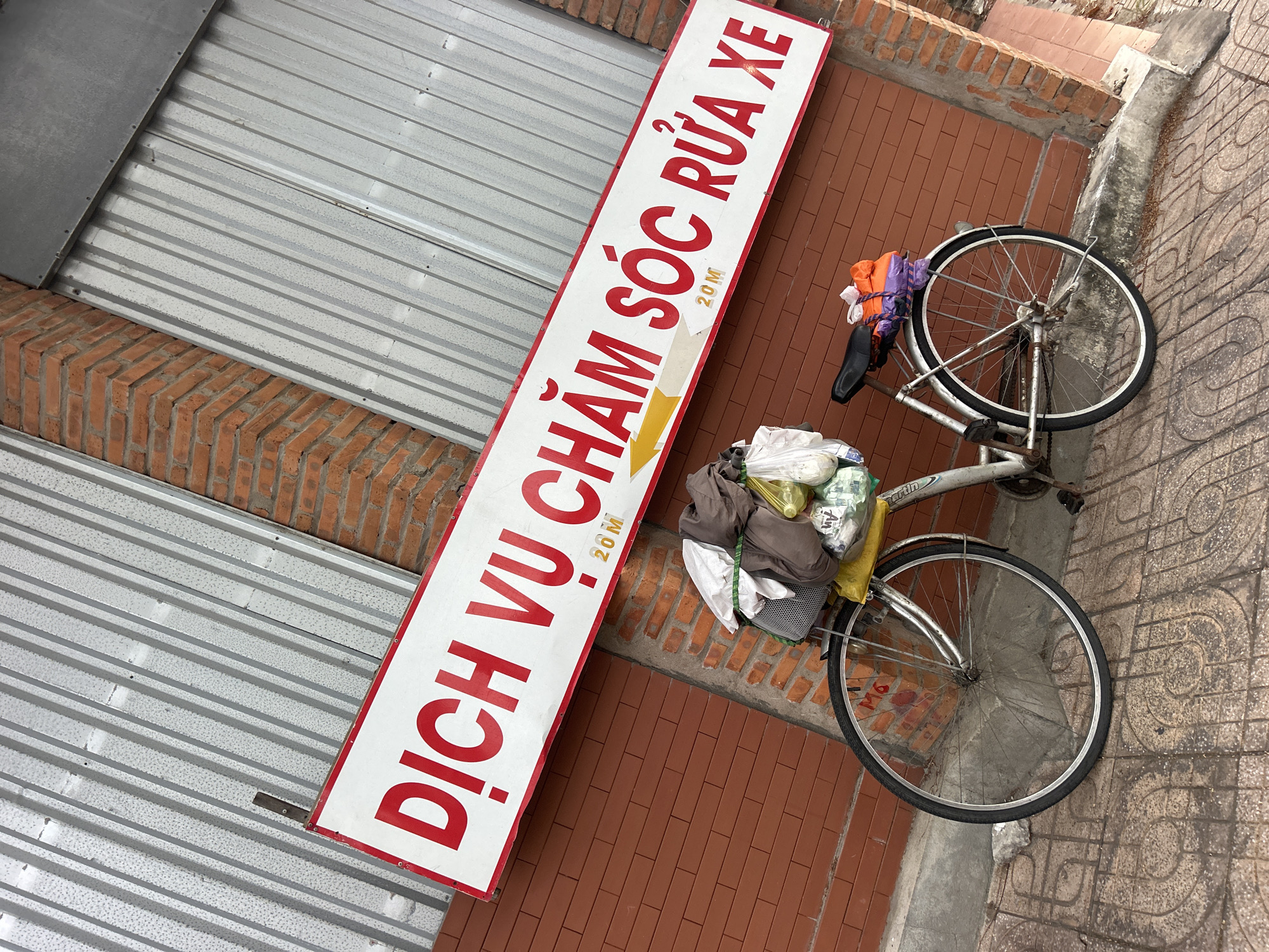 A sign in Vietnamese reading "Dịch Vụ Chăm Sóc Rửa Xe" with an arrow pointing right and the distance "20M," indicating a car wash or vehicle care service, is propped up next to a gray corrugated metal wall and a red brick wall in Nha Trang Ward, Vietnam. A bicycle loaded with various bags is parked nearby on a patterned sidewalk and rests against the brick wall.