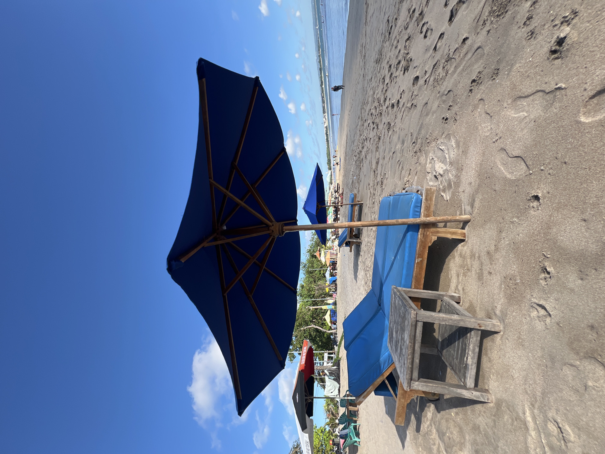The image shows a beach scene in Kuta, Indonesia. The foreground features a blue beach umbrella and a wooden lounge chair with blue cushions, positioned on a sandy beach. Further down the beach, there are more umbrellas and chairs. In the distance, the ocean is visible, with a person standing in the water. The sky is blue with some scattered clouds.