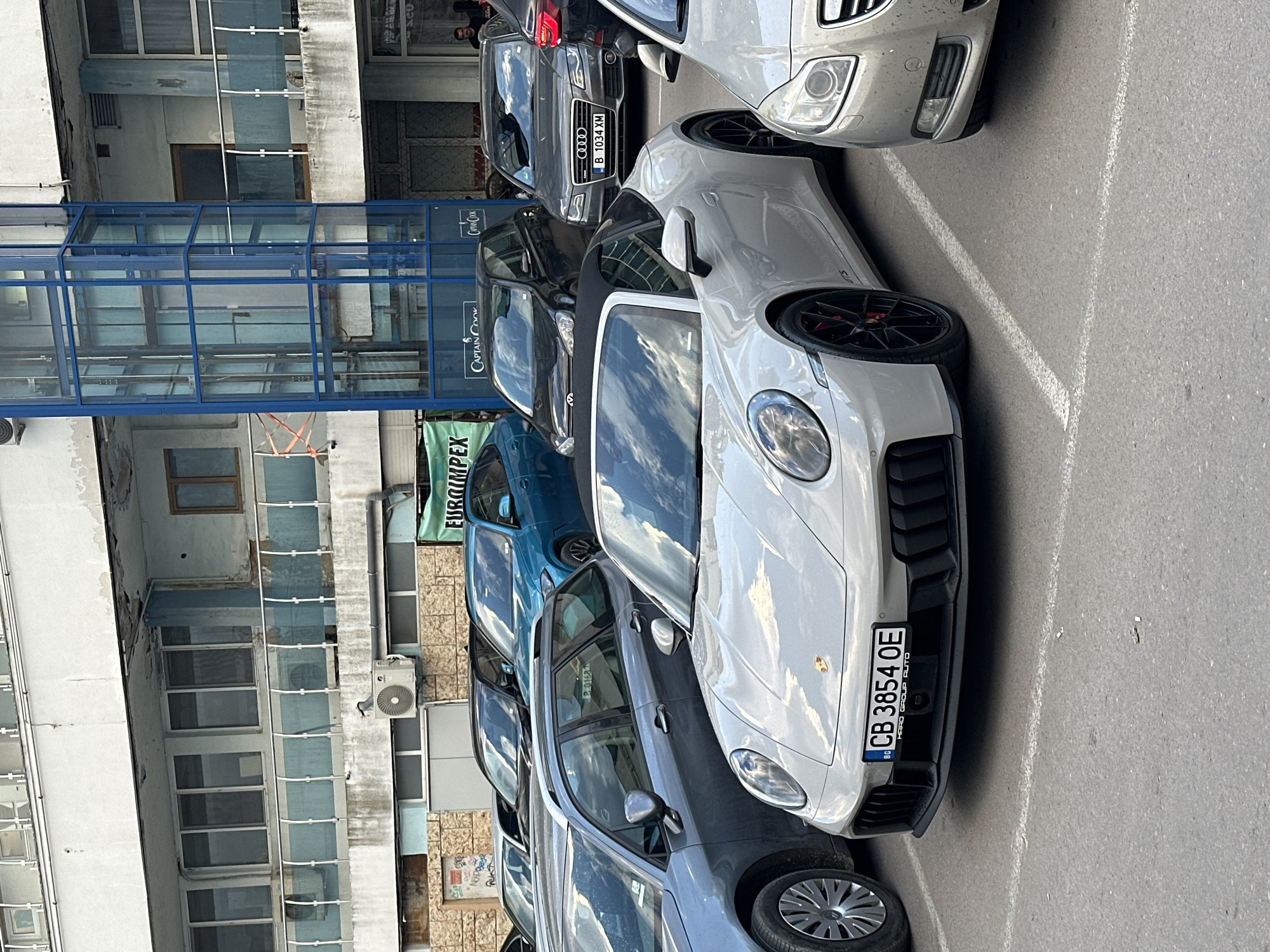 The image shows a street parking scene, likely in an urban area. Several cars are parked in a row, including a white Porsche with a Bulgarian license plate "CB 3854 OE," a gray Audi with the license plate "B 1034 XM," and a blue car with a partial plate "P 0168" visible.
The background features a multi-story building with a glass-enclosed walkway bearing the name "CAPTAIN COOK". There is a sign for "EUROIMPEX" visible further back. This location is likely in Varna, Bulgaria, given the Bulgarian license plates. A couple of individuals can be seen in the background near the Audi.