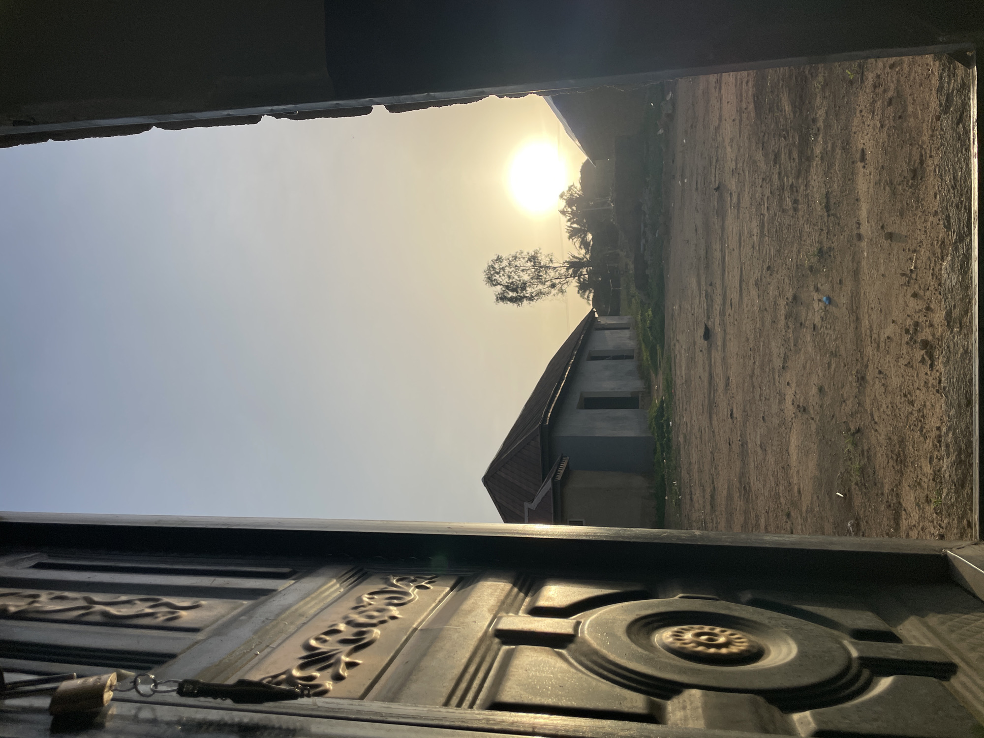 The image depicts a view through a doorway or opening. In the distance, a building with a pitched roof stands in a sunlit clearing. A lone tree rises near the building. The foreground is dominated by a detailed, dark-colored metal door, partially obscuring the view. The location is likely Durumi, Nigeria. The scene is bathed in bright, hazy sunlight. No people are visible.