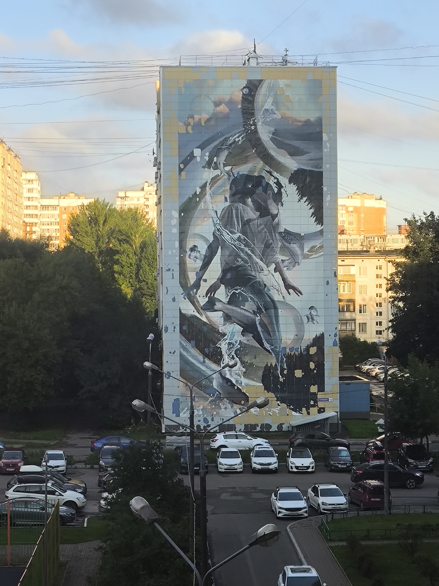 An exterior, eye-level shot captures a large mural painted on the side of a multi-story apartment building. The mural depicts a stylized human figure in a dynamic pose, surrounded by water and fish elements, rendered in a mix of blues, grays, and whites. The building itself is constructed of light-colored bricks and has a grid of windows. In front of the building, a street is visible with parked cars and streetlights. Trees and other apartment buildings are seen in the background, with a cloudy sky overhead. The overall scene suggests an urban residential area.