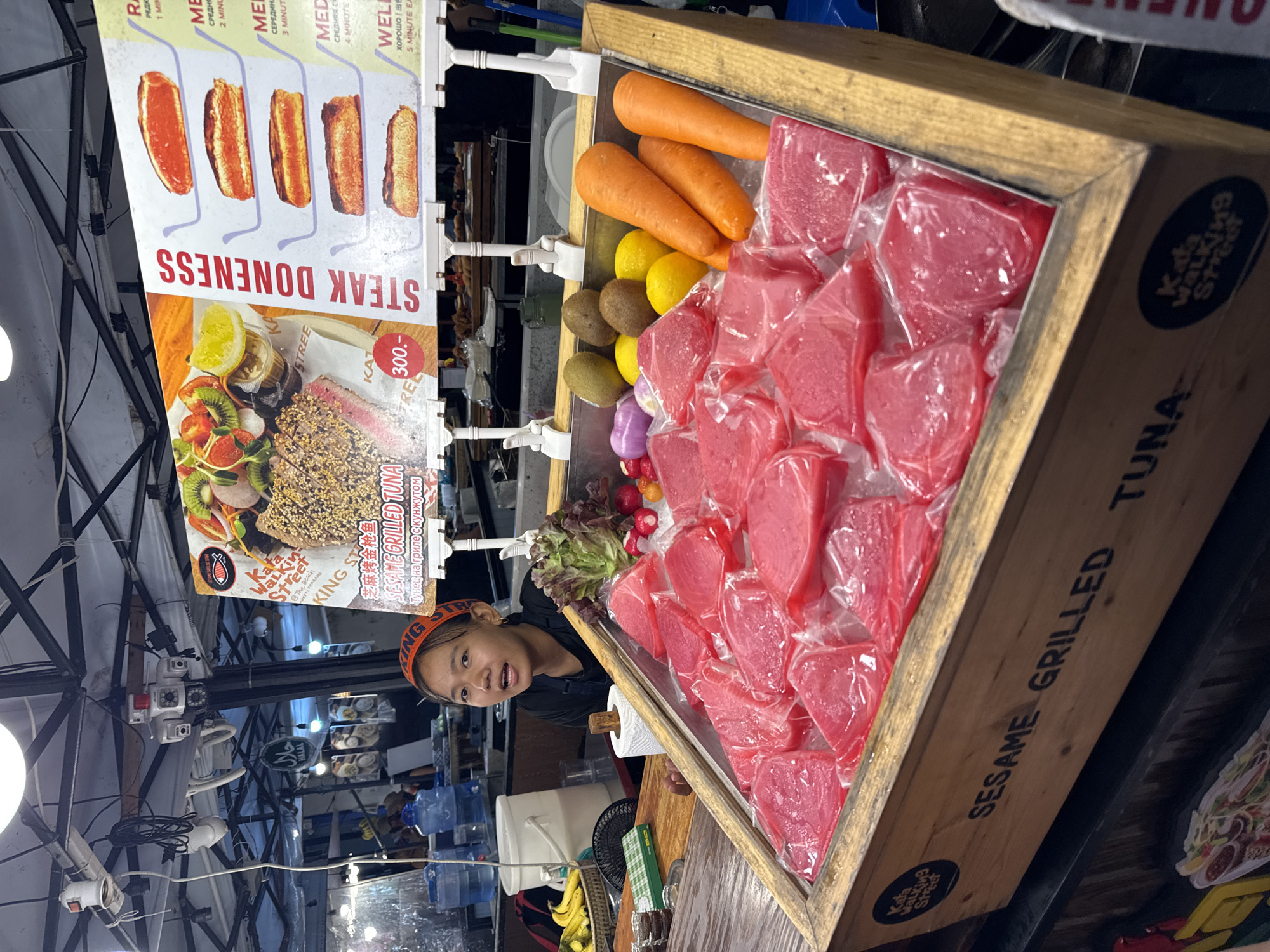 In a bustling street market, likely in Karon, Thailand, a young woman with an orange headband is featured prominently, looking towards the camera with a slight smile. She is positioned behind a display of food items.
The foreground is dominated by a wooden crate filled with an abundance of bright red, raw tuna steaks, each individually packaged in plastic. Adjacent to the tuna, in a separate section of the display, are fresh produce including lemons, kiwis, carrots, and leafy greens. A small sign indicates a price of "300.-" in what appears to be Thai Baht.
Above the food display, a colorful menu board is partially visible, advertising "STEAK DONENESS" with images depicting different levels of steak doneness, ranging from rare to well-done. The text on the board includes various languages, suggesting a tourist-heavy location.
In the background, there are glimpses of other market stalls, water dispensers, and electrical wiring, indicative of an outdoor market setting. A "HALAL" sign is also visible, suggesting a diverse range of food offerings. The overall impression is a lively and vibrant food market with a focus on fresh ingredients and diverse culinary options. The lighting suggests it could be daytime or early evening.