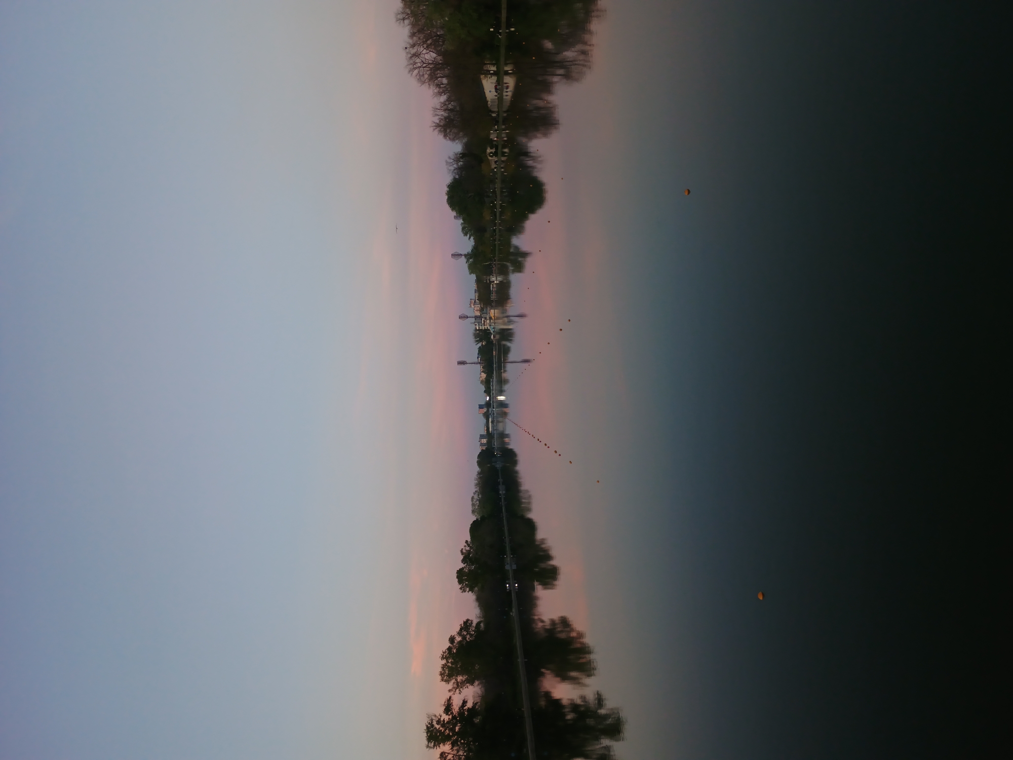 The image, presented vertically, captures a twilight landscape over a body of water, likely a river or canal in Plovdiv, Bulgaria, with a prominent reflection. The left side of the image features the actual sky, displaying a gentle gradient from pale blue-gray at the top to soft pink and orange hues towards the bottom, indicative of either sunrise or sunset. A small bird is seen in flight against the sky in the upper left corner.
Horizontally bisecting the image is a narrow strip of land. This strip is densely lined with dark, silhouetted trees and contains visible structures resembling streetlights or small buildings.
The right side of the image depicts the calm surface of the water, which creates a strong, mirror-like reflection of the sky, trees, and structures. The reflected sky colors mirror those on the left, transitioning into darker, muted tones further to the right. Scattered across the water's surface, particularly in the lower right, are several small, bright orange or yellow spherical objects, possibly floating lanterns or decorative buoys. A long, faint line of dark, small objects, likely buoys or markers, also extends horizontally across the reflective water surface. The still water and soft, dim lighting contribute to a serene and tranquil atmosphere. No discernible text is present.