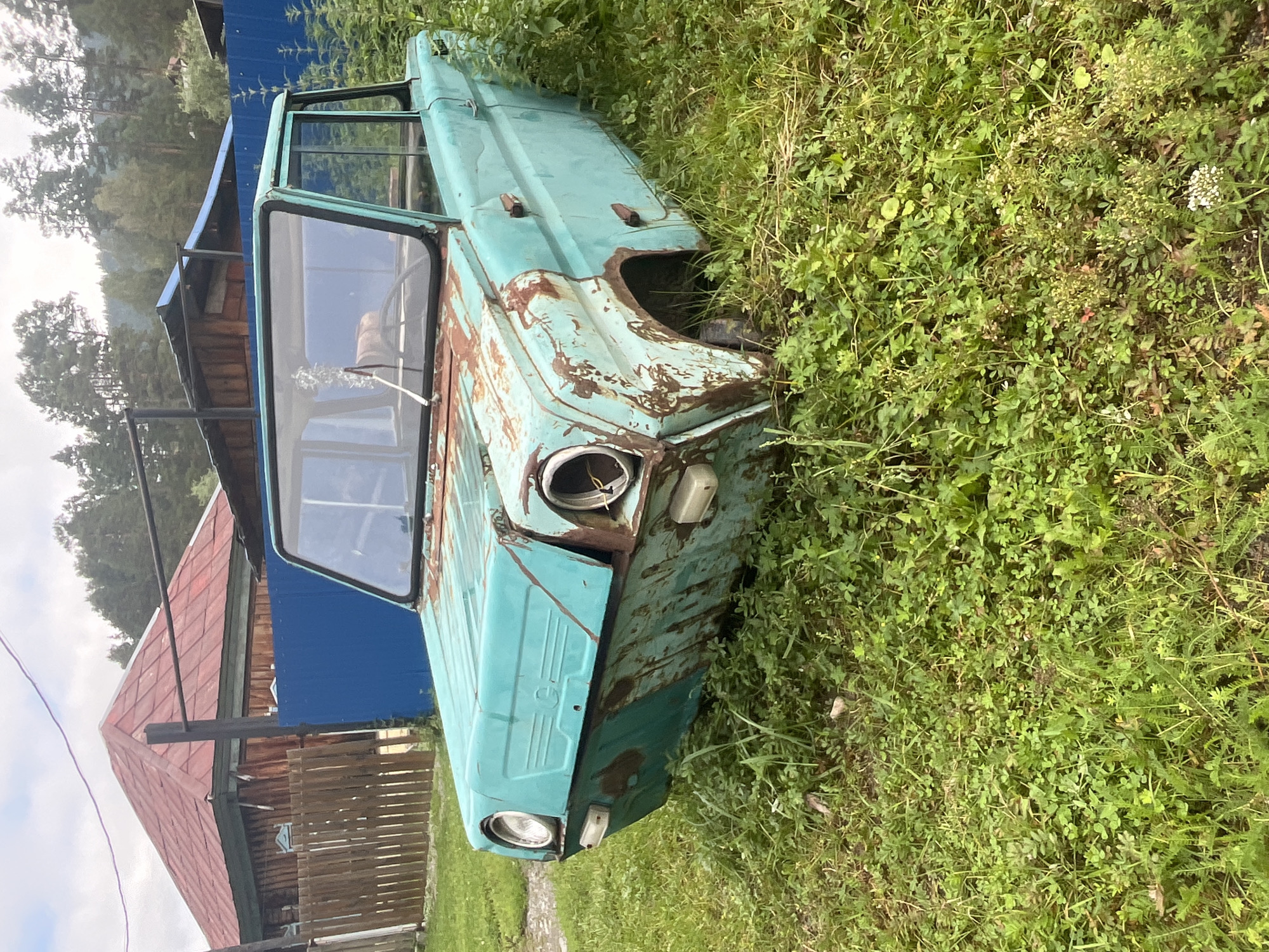 An abandoned, turquoise, vintage amphibious vehicle is parked in a grassy yard in Ust-Sema, Russia. The vehicle, which appears to be in a state of disrepair, has significant rust and missing parts, including its wheels. The car is surrounded by overgrown green grass and weeds. In the background, a wooden house with a red roof is visible, along with a blue fence and trees. The sky is overcast.
