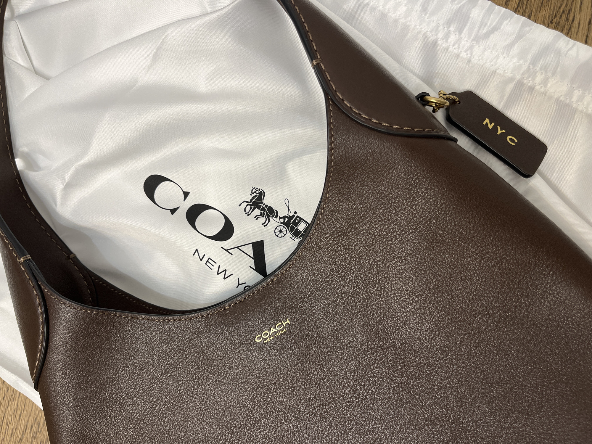 A brown leather Coach handbag is partially visible, lying on a white cloth bag with the Coach logo printed on it in black. The handbag has a gold logo embossed on its surface that reads "COACH NEW YORK". A dark brown leather tag with "NYC" embossed in gold hangs from the handbag. The scene appears to be indoors, possibly on a wooden surface, with no indication of the city of Limassol, Cyprus. There are no people present in the image.