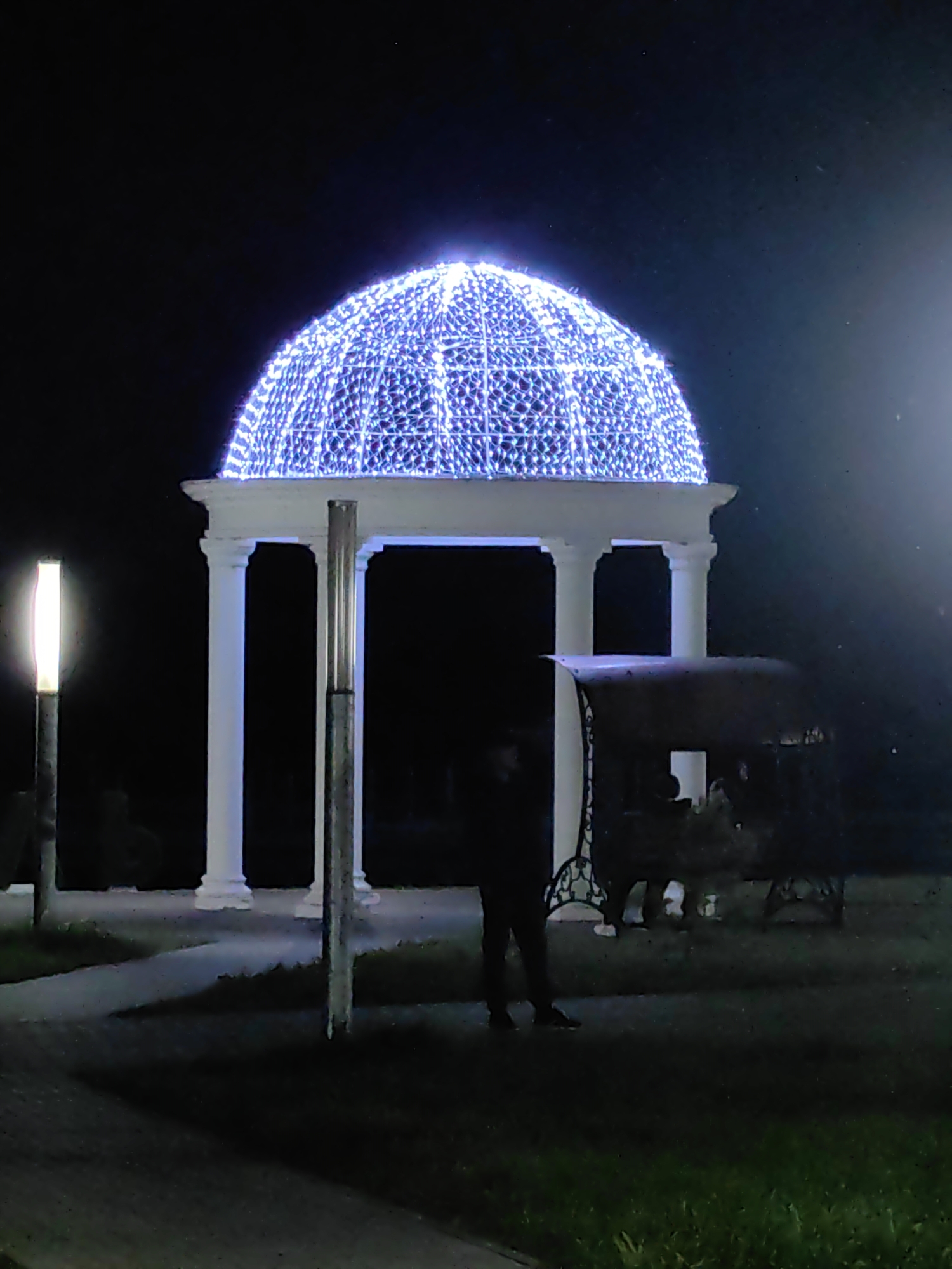 The image depicts a night scene in Myadzyel, Belarus, featuring a white gazebo with a dome covered in blue lights. The gazebo has four white columns and appears to be a focal point in an outdoor area. In the foreground, to the left of the gazebo, is a tall, illuminated post with a light fixture. To the right of the gazebo, there is a dark, ornate bench or seating structure. A person, silhouetted against the background and illuminated by the lights, stands near the bench. The ground is paved, and grass is visible in the lower right portion of the image. The overall atmosphere is dark, with the blue lights on the gazebo providing a prominent source of illumination.