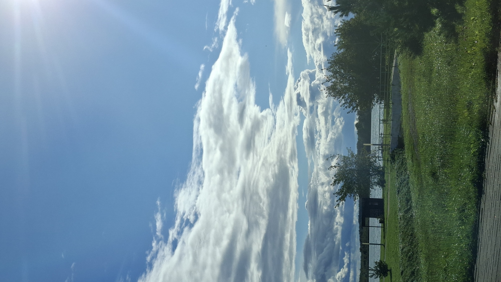 A bright, sunny day with a vast blue sky filled with fluffy white and grey clouds. The sun is visible in the upper right corner, casting a bright glare. Below the sky, a calm body of water, likely a lake, stretches across the horizon, bordered by a distant treeline. In the foreground, a grassy park area slopes down towards the water. There are several trees scattered throughout the park, with a small dark structure, possibly a shelter or shed, located near the water's edge. A walkway or path is visible in the lower foreground. The scene is serene and suggests an outdoor recreational area. This image depicts a view of the Myadzyel Lake in Myadzyel, Belarus.