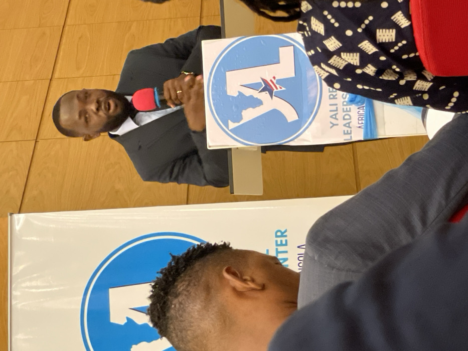 An indoor scene captures a presentation or discussion at a YALI (Young African Leaders Initiative) event in Luanda, Angola. The main subject is a man of African descent, dressed in a dark suit and white shirt, speaking animatedly into a red and black microphone. He has a short beard and mustache, and a ring is visible on his left hand.
Next to the speaker, a light blue and white circular sign displays the YALI logo, which includes a map of Africa and a star. Text on a banner beneath this sign reads "YALI REGIONAL LEADERS CENTER AFRICA," though parts are obscured. Another person, with dark, patterned attire and a red shoulder strap, is partially visible seated to the speaker's right.
In the foreground, the back of another individual's head, featuring short, curly hair, suggests an audience member. A large banner, similar to the one beside the speaker, is also visible in the lower part of the frame, with partially legible text including "CENTER" and "ANGOLA," further indicating the event's location and nature. The background consists of light wooden wall paneling. The scene conveys an atmosphere of engagement and leadership development.