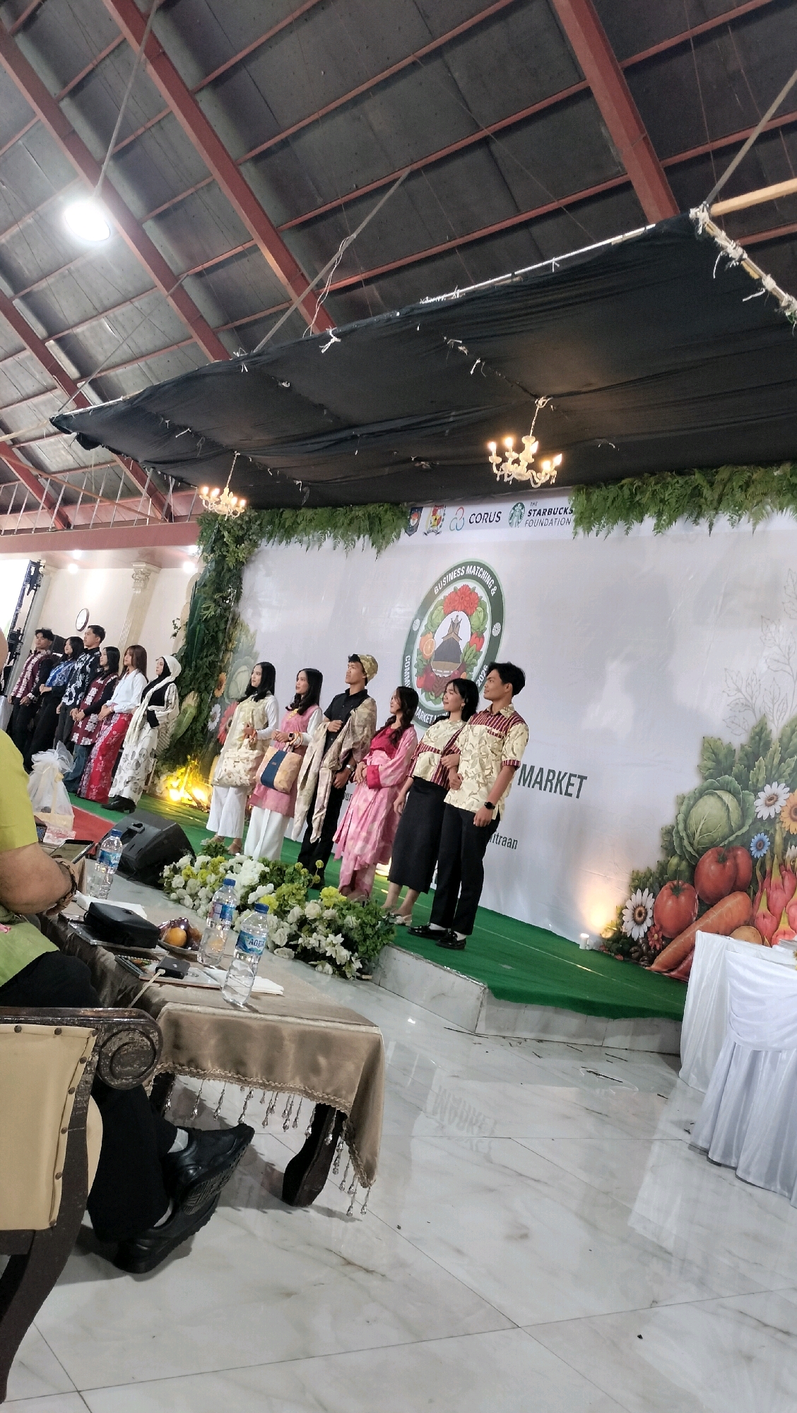 In Durin Serugun, Indonesia, a group of young adults, likely students, pose on a stage at what appears to be an indoor event. They are dressed in traditional attire, with some individuals wearing batik-patterned clothing. The stage is decorated with green foliage and a large banner that reads "BUSINESS MATCHING & COMMUNITY MARKET 2023." The backdrop features various logos, including "CORUS" and "THE STARBUCKS FOUNDATION." In the foreground, a table covered with a brown tablecloth and adorned with fringe and decorative beads is visible. Several bottles of water and some fruit are placed on the table. The floor is tiled with a marble pattern. The event appears to be held in a large hall with a high ceiling supported by visible beams and illuminated by chandeliers.